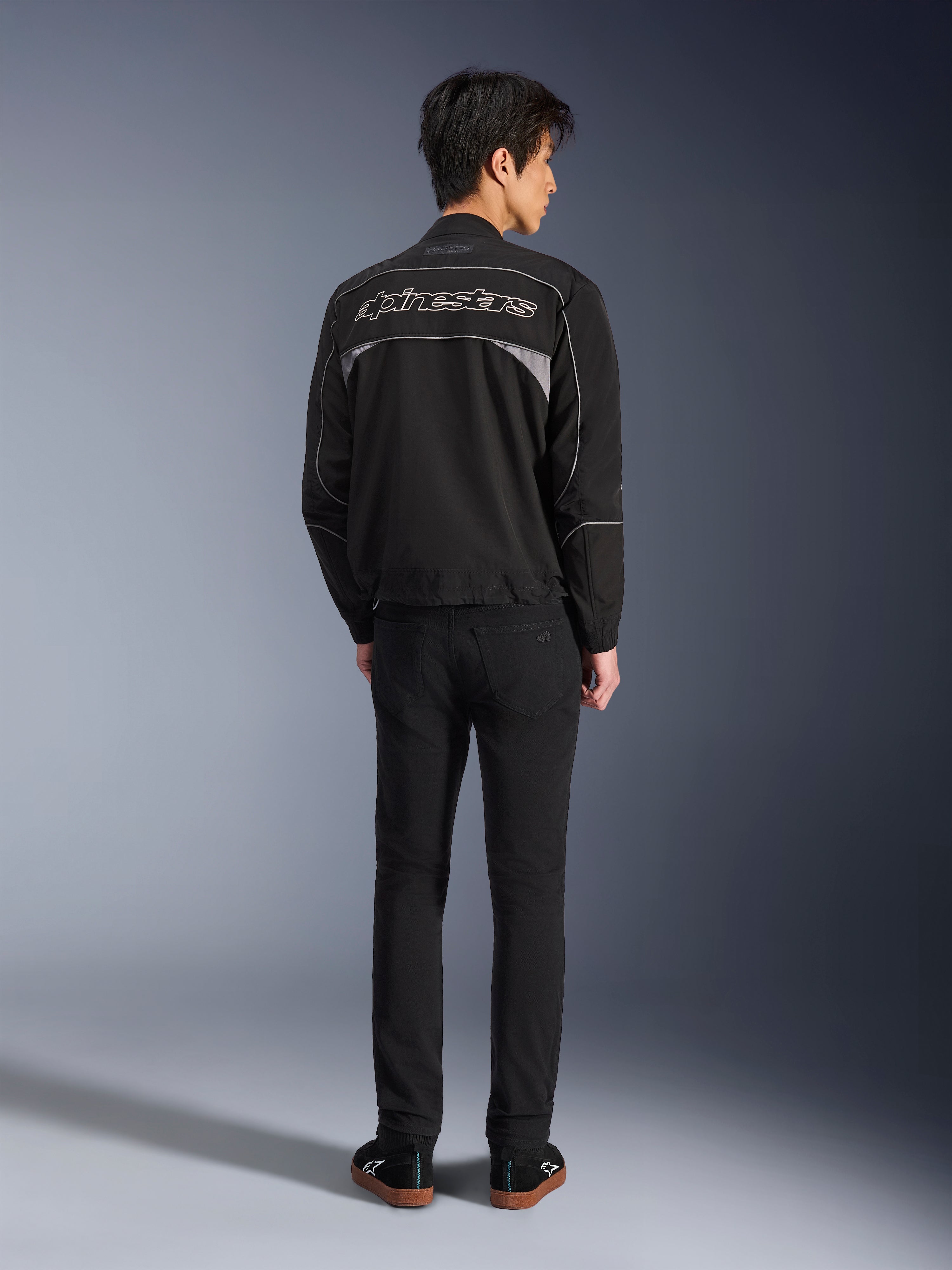 Alpinestars Unite Jakke, sort og mørkegrå motorcykeljakke, set bagfra med Alpinestars outline-logo på ryggen, grå reflekterende paneler, minimalistisk urbant design, båret med sorte bukser og sorte sko