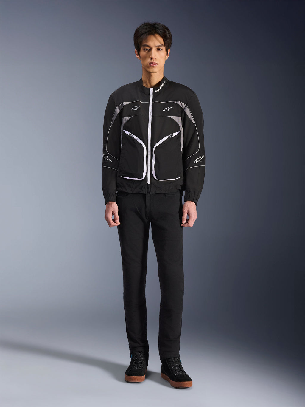 Alpinestars Unite-jakke, motorcykeljakke, sort og mørkegrå, båret af en model med sorte bukser og sorte støvler, med hvide kontrastlynlåse, reflekterende piping og minimalistisk urbant design