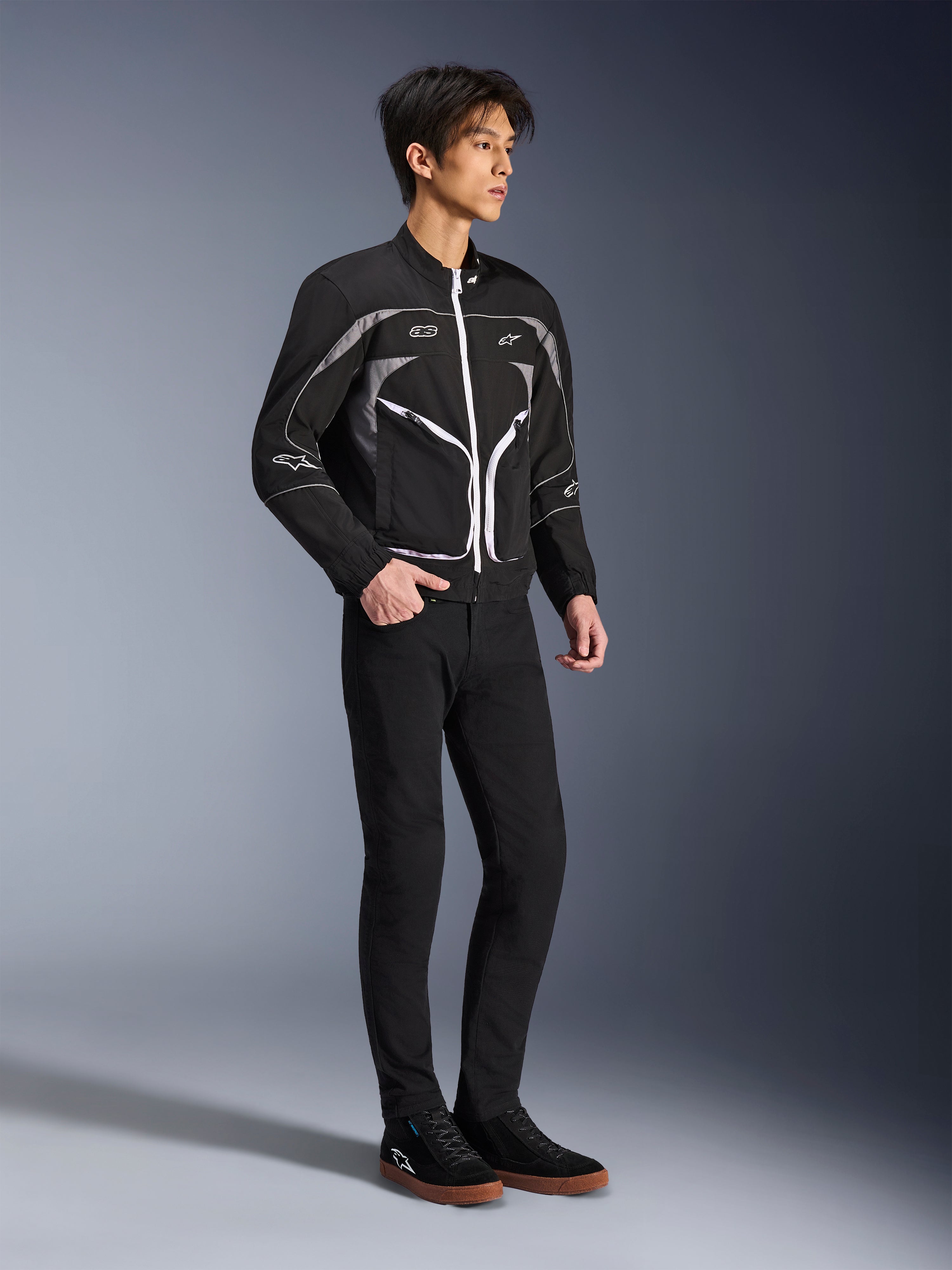 Model iført Alpinestars Unite-jakke i sort og mørkegrå, urban motorcykeljakke med hvide lynlåse og reflekterende detaljer, kombineret med sorte slim-fit bukser og sneakers, minimalistisk gadeinspireret køreudstyr.