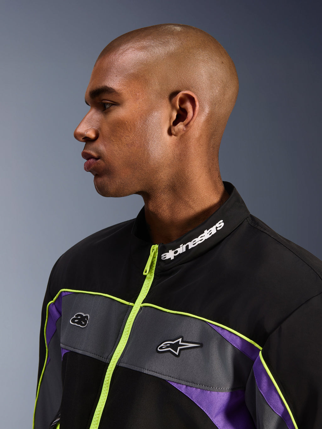 Alpinestars Unite-jakke, Black Asphalt Violet, profilvisning af mandlig model med fokus på den høje krave med hvid Alpinestars-branding, neongul lynlås, grå brystpaneler med logoer, lilla detaljer, minimalistisk urban motorcykel-streetwear-styling