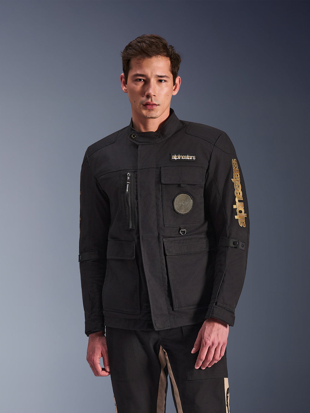 Alpinestars Dice Jacket, sort motorcykeljakke, båret af model, med guld Alpinestars-logoer, flere funktionelle lommer, cirkulært spiralmærke og urban feltjakke-styling