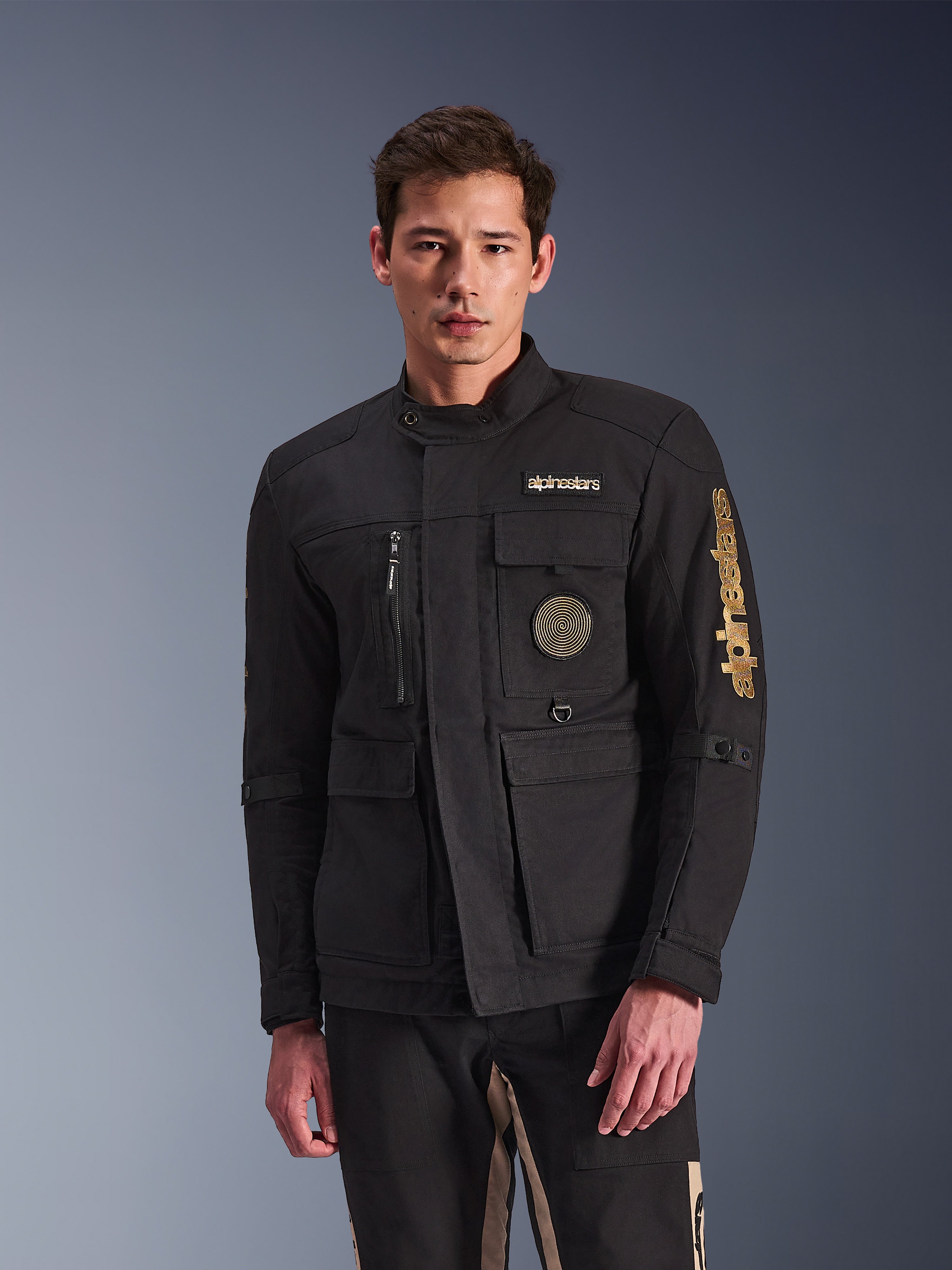 Alpinestars Dice Jacket, sort motorcykeljakke, båret af model, med guld Alpinestars-logoer, flere funktionelle lommer, cirkulært spiralmærke og urban feltjakke-styling
