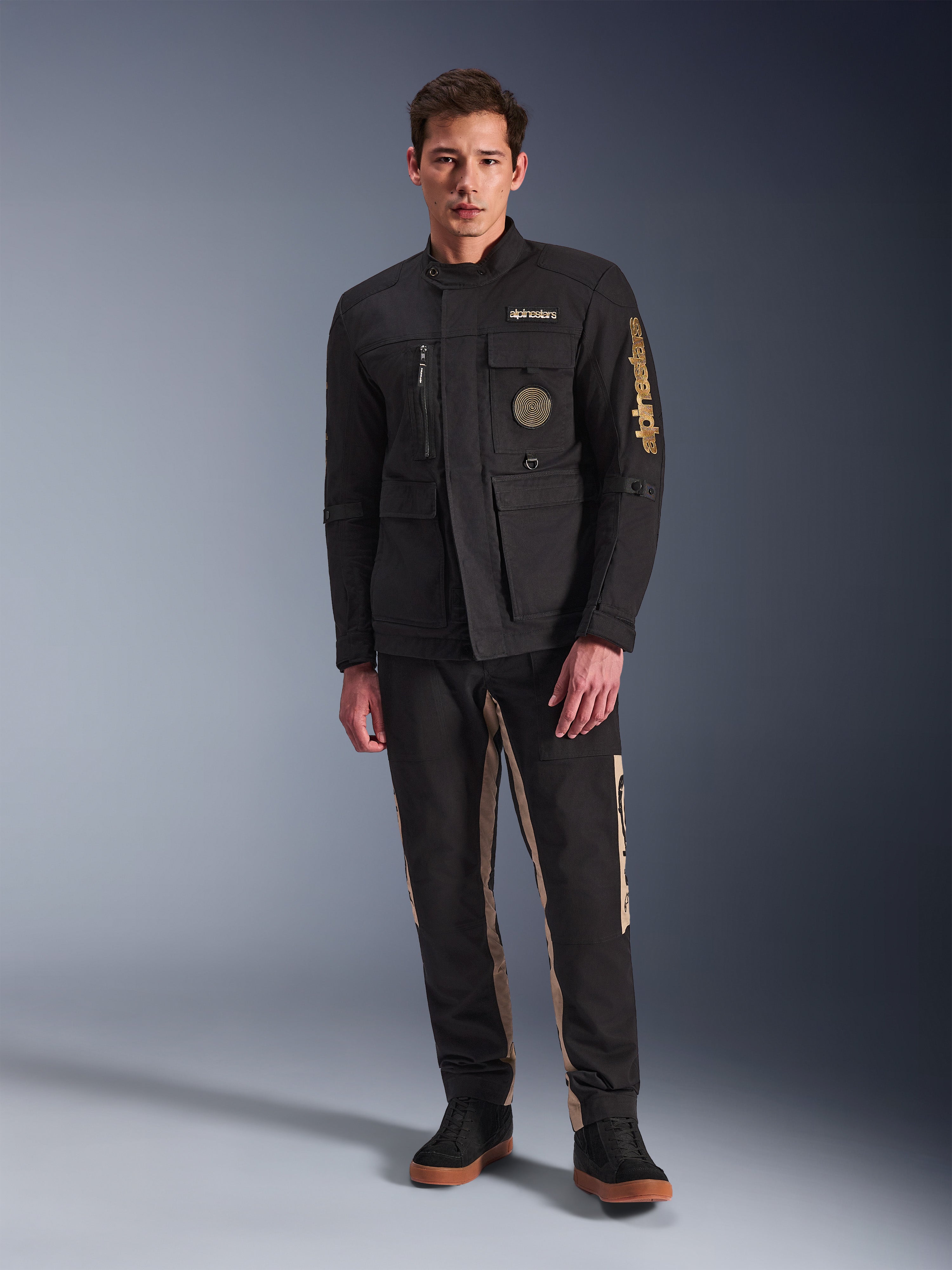 Mandlig model iført Alpinestars Dice Jacket, sort urban motorcykeljakke med vintage feltjakke-stil, fire funktionelle frontlommer, Alpinestars-branding på ærmet og brystet, kombineret med sorte og sandfarvede bukser, moderne urban køreudstyr