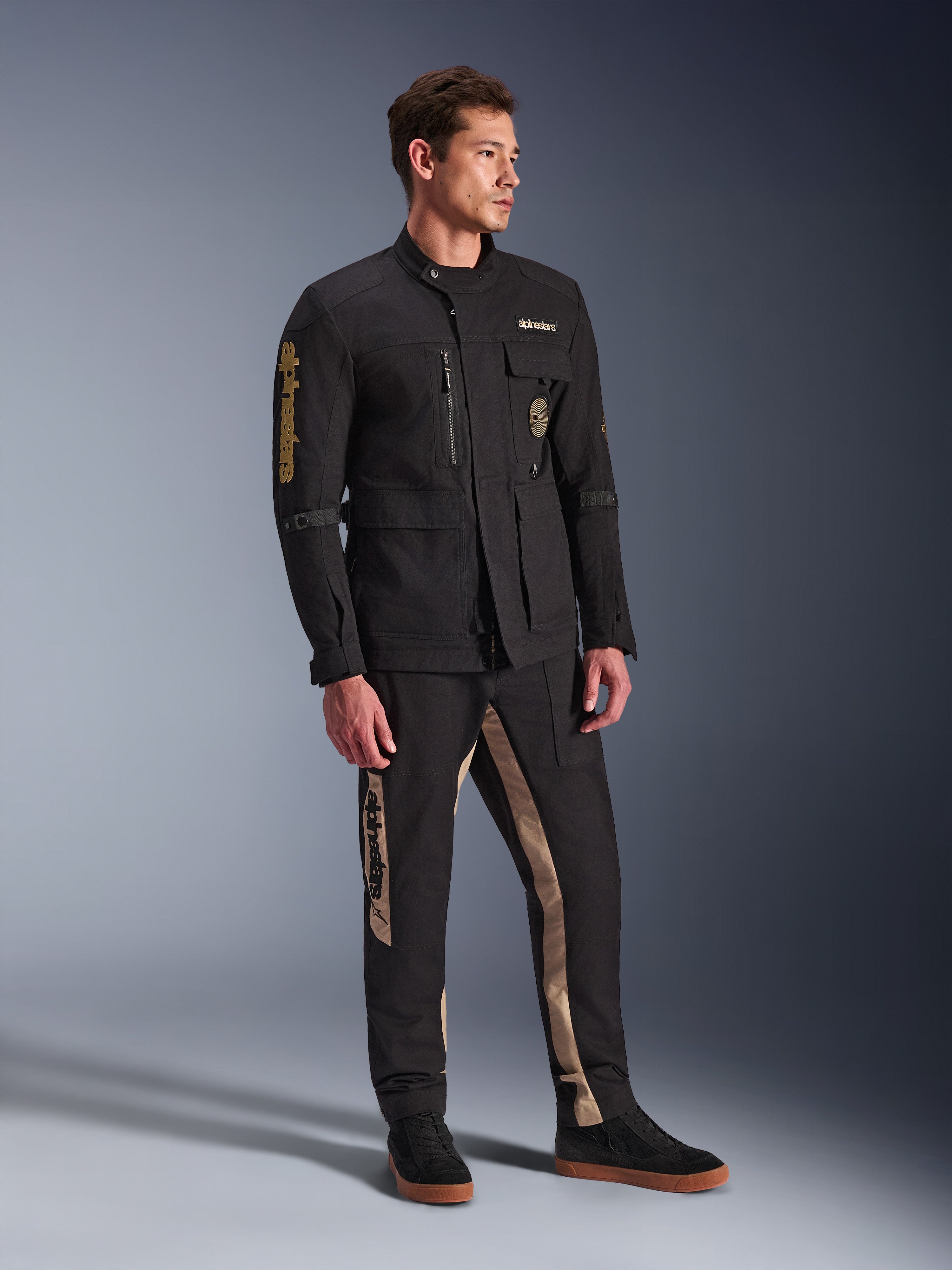 Mandlig model iført Alpinestars Dice-jakke i sort, urban motorcykeljakke med vintage feltjakke-æstetik, med flere funktionelle lommer og guld-branding på ærmet, kombineret med matchende sorte og sandfarvede bukser og sorte sneakers.