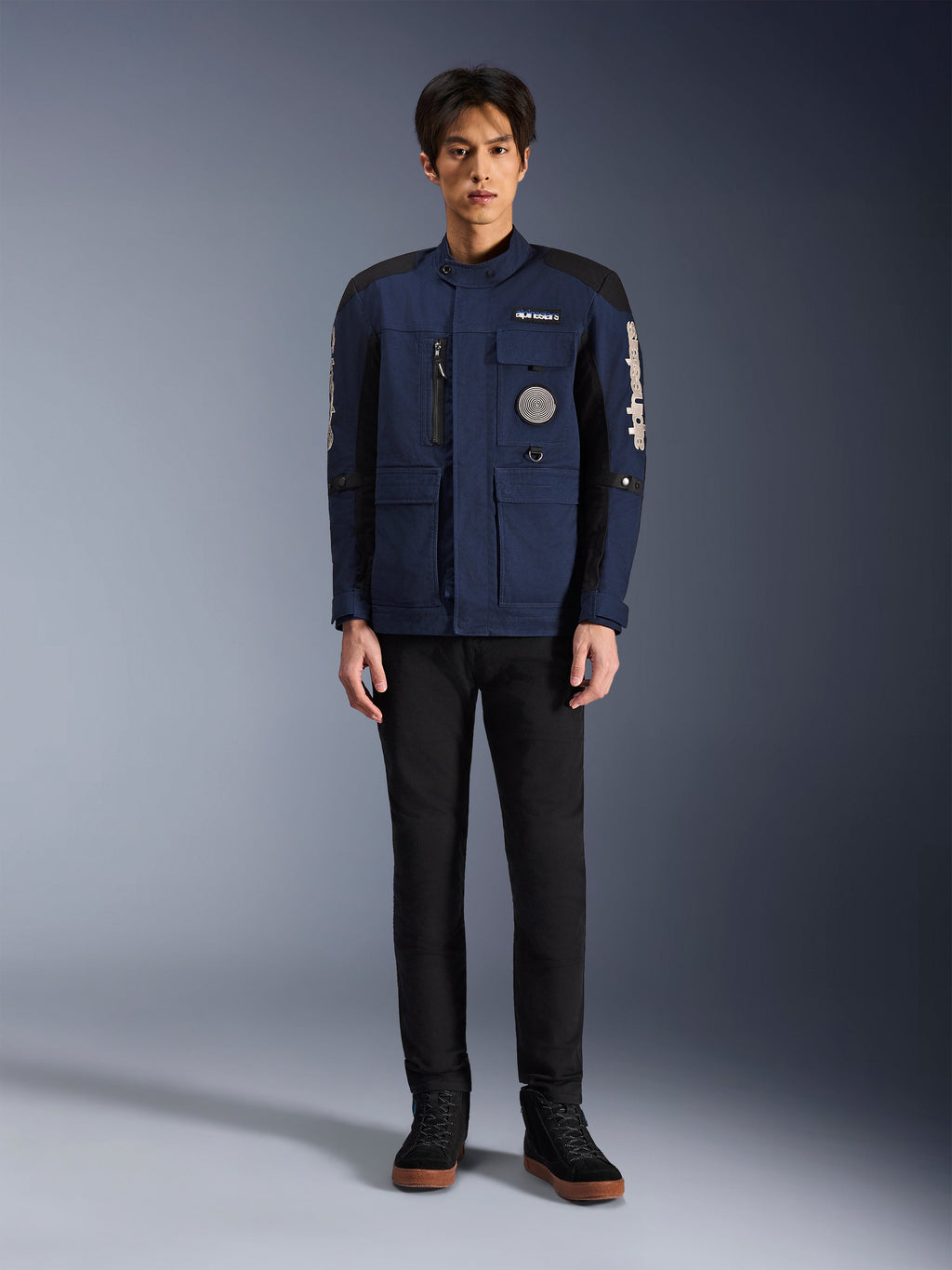 Alpinestars Dice-jakke, urban motorcykeljakke, mørkeblå og sort, bæres af en model med sorte bukser og sorte sko, moderne feltjakke-design med flere lommer, lodret brystlynlås og hvidt Alpinestars-branding på ærmerne.