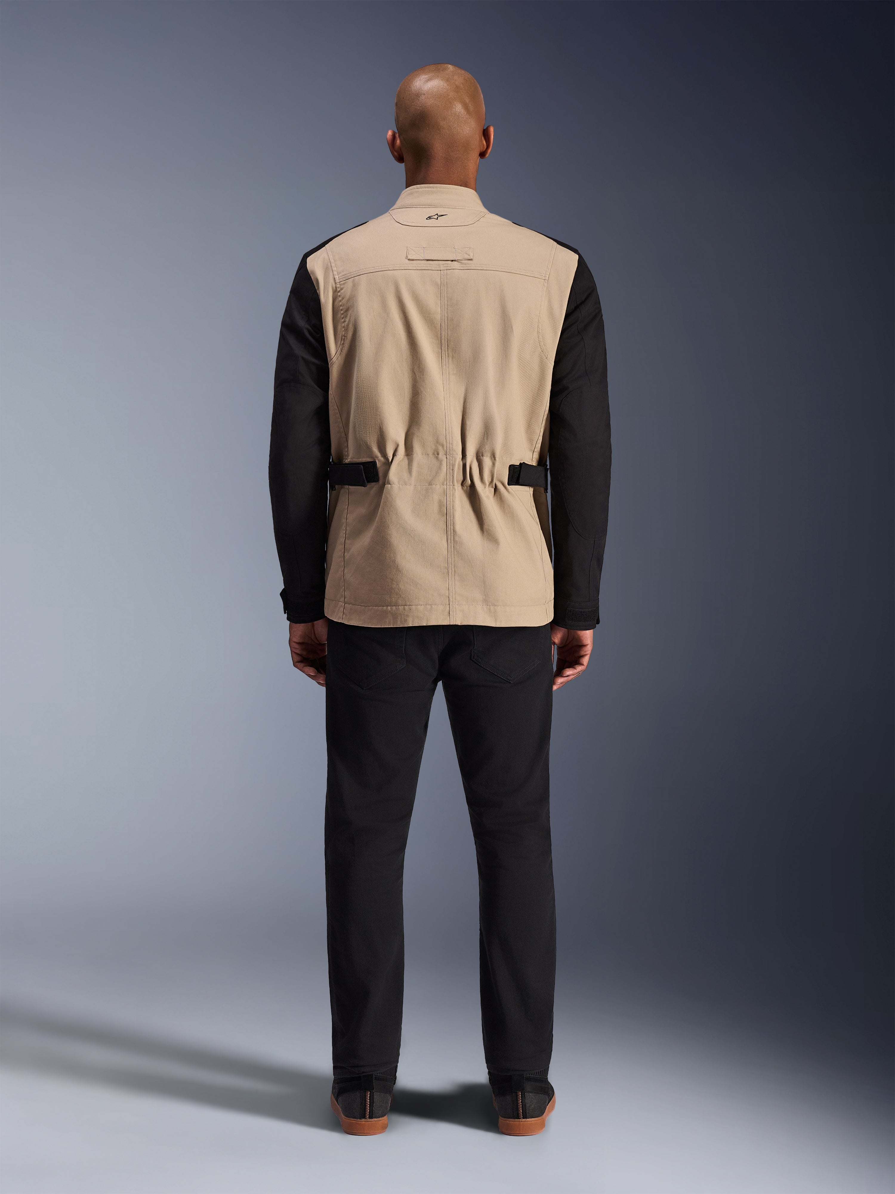 Alpinestars Dice-jakke, urban motorcykeljakke, tan og sort, set bagfra på model, med polybomuld twill-konstruktion, justerbare taljestropper og lille broderet logo på kraven, båret med sorte bukser.