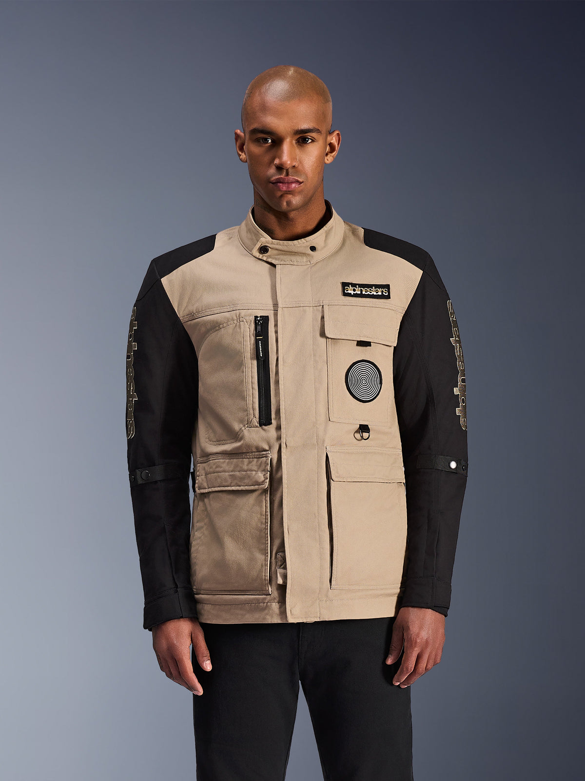Alpinestars Dice Jacket, urban motorcykeljakke, beige og sort, robust arbejdstøjsæstetik med flere brugslommer og brystpatches, høj krave med knaplukning, markant branding på ærmerne, båret af en mandlig model