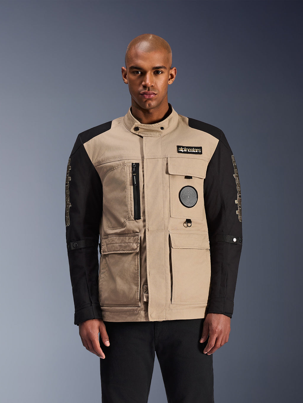 Alpinestars Dice Jacket, urban motorcykeljakke, beige og sort, robust arbejdstøjsæstetik med flere brugslommer og brystpatches, høj krave med knaplukning, markant branding på ærmerne, båret af en mandlig model
