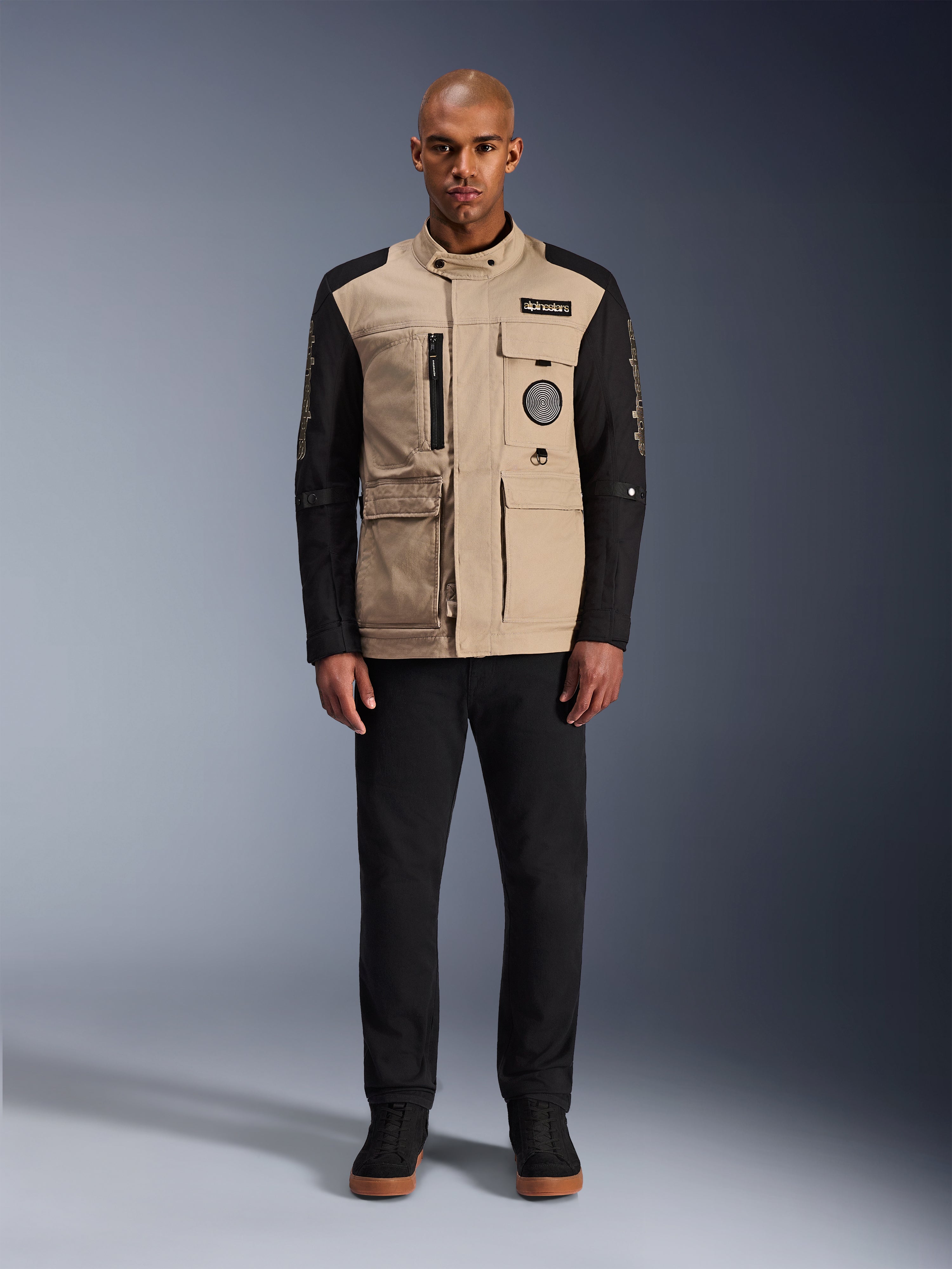 Alpinestars Dice-jakke, urban motorcykeljakke, aluminiums-tan krop med sorte ærmer, arbejdstøjsinspireret silhuet med fire funktionelle forlommer, bæres med sorte bukser og sorte high-top sneakers, robust og gadeklart køreudstyr