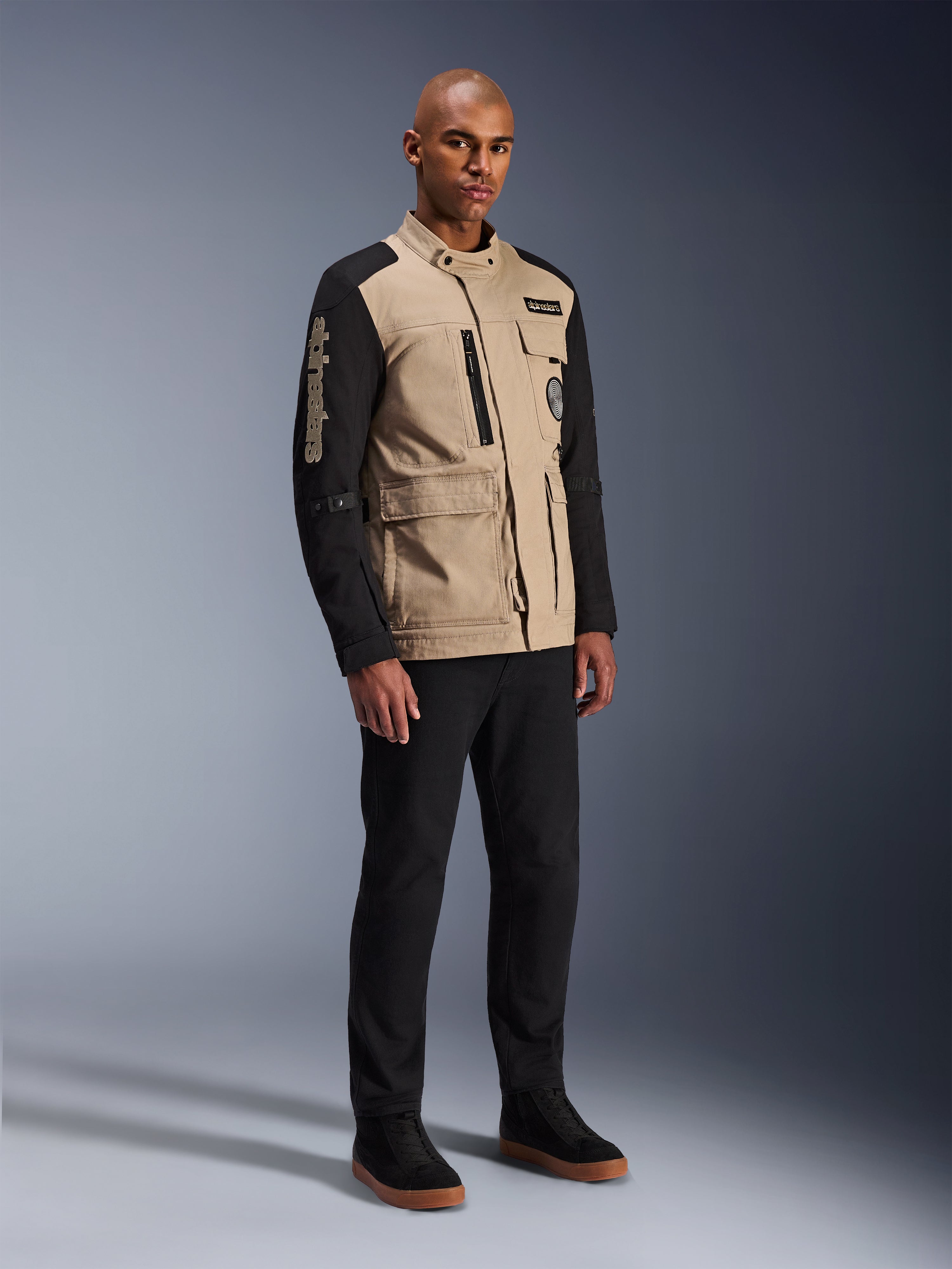 Alpinestars Dice-jakke, urban motorcykeljakke, Aluminum Black og tan, tofarvet design med sorte ærmer og khaki torso, båret af en model med sorte bukser og sorte sneakers, arbejdstøjsinspireret æstetik med funktionelle lommer og broderet branding på ærmet.