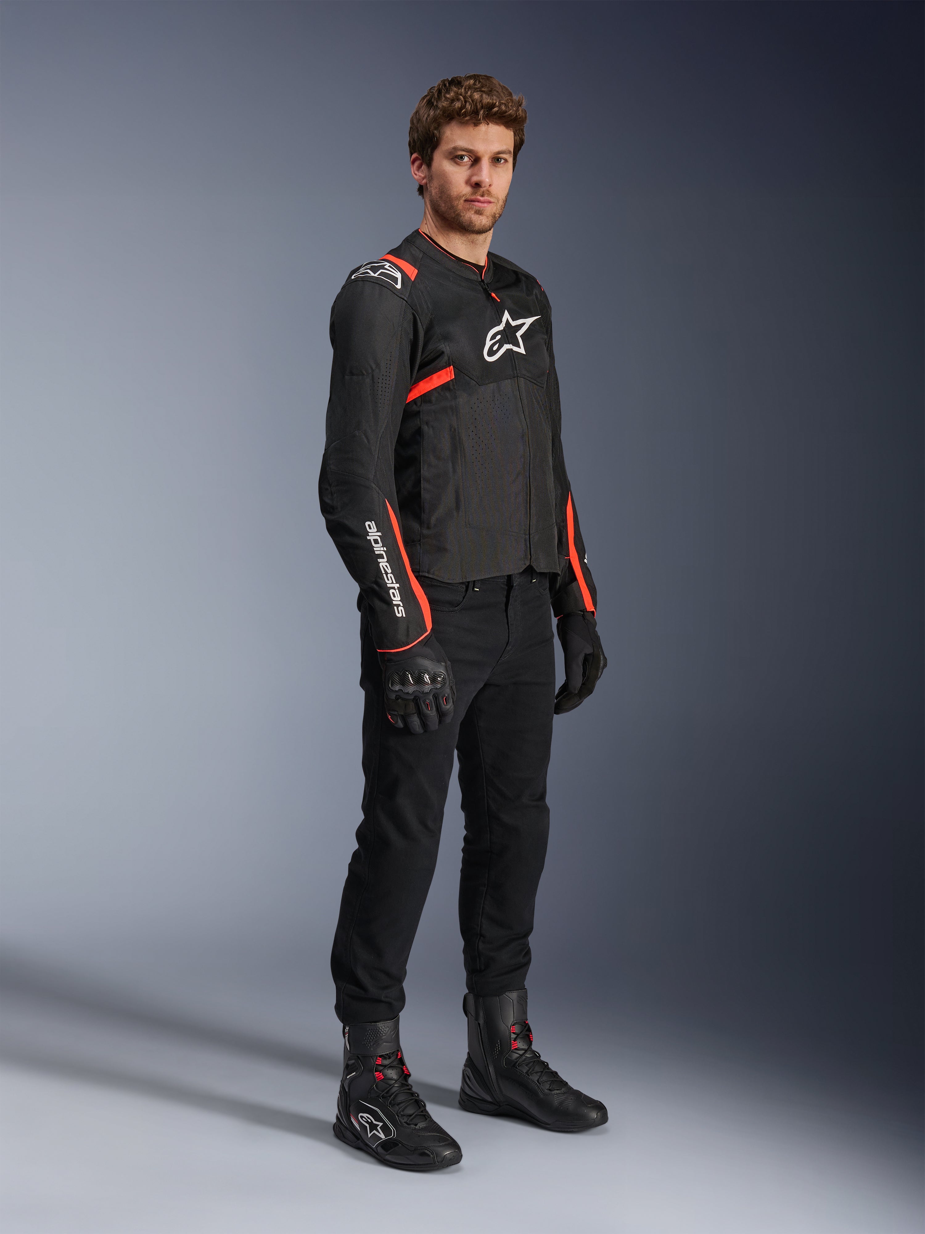 Male model wearing Alpinestars T-Sps Air V2 Jacket, Black with Red Fluo accents, paired with black motorcycle pants, black gloves, and black boots, highlighting a sporty design with mesh panels and white Alpinestars logos.
