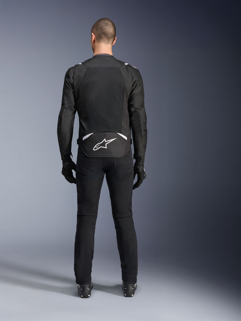 Alpinestars T-Sps Air V2 Jakke, set bagfra, sort og hvid tekstil motorcykeljakke, med et stort mesh-panel på ryggen for maksimal luftgennemstrømning og hvidt Astars-logo på den nederste del af ryggen, båret med sorte motorcykelbukser og handsker