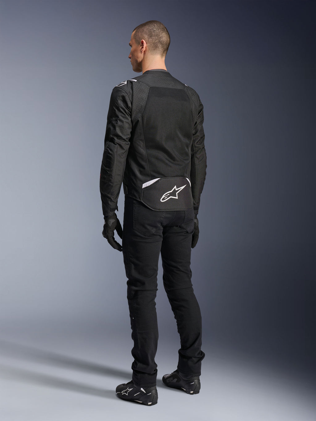 Alpinestars T-Sps Air V2 Jakke, tekstil motorcykeljakke, sort, set bagfra med et stort mesh-ventilationspanel og hvidt Alpinestars-logo nederst på ryggen, kombineret med sorte kørebukser og handsker for et sporty urbant look.