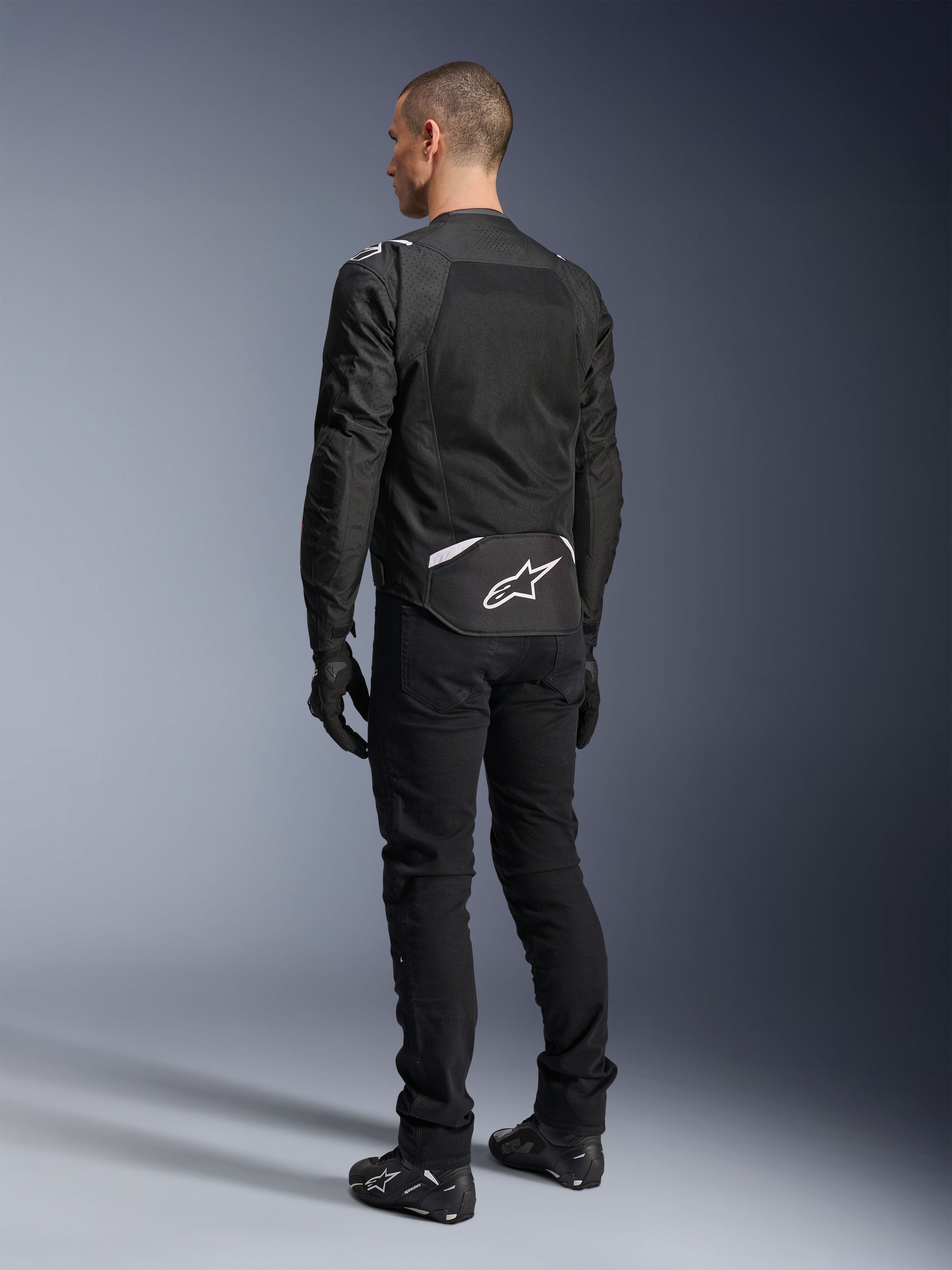 Alpinestars T-Sps Air V2 Jakke, tekstil motorcykeljakke, sort, set bagfra med et stort mesh-ventilationspanel og hvidt Alpinestars-logo nederst på ryggen, kombineret med sorte kørebukser og handsker for et sporty urbant look.