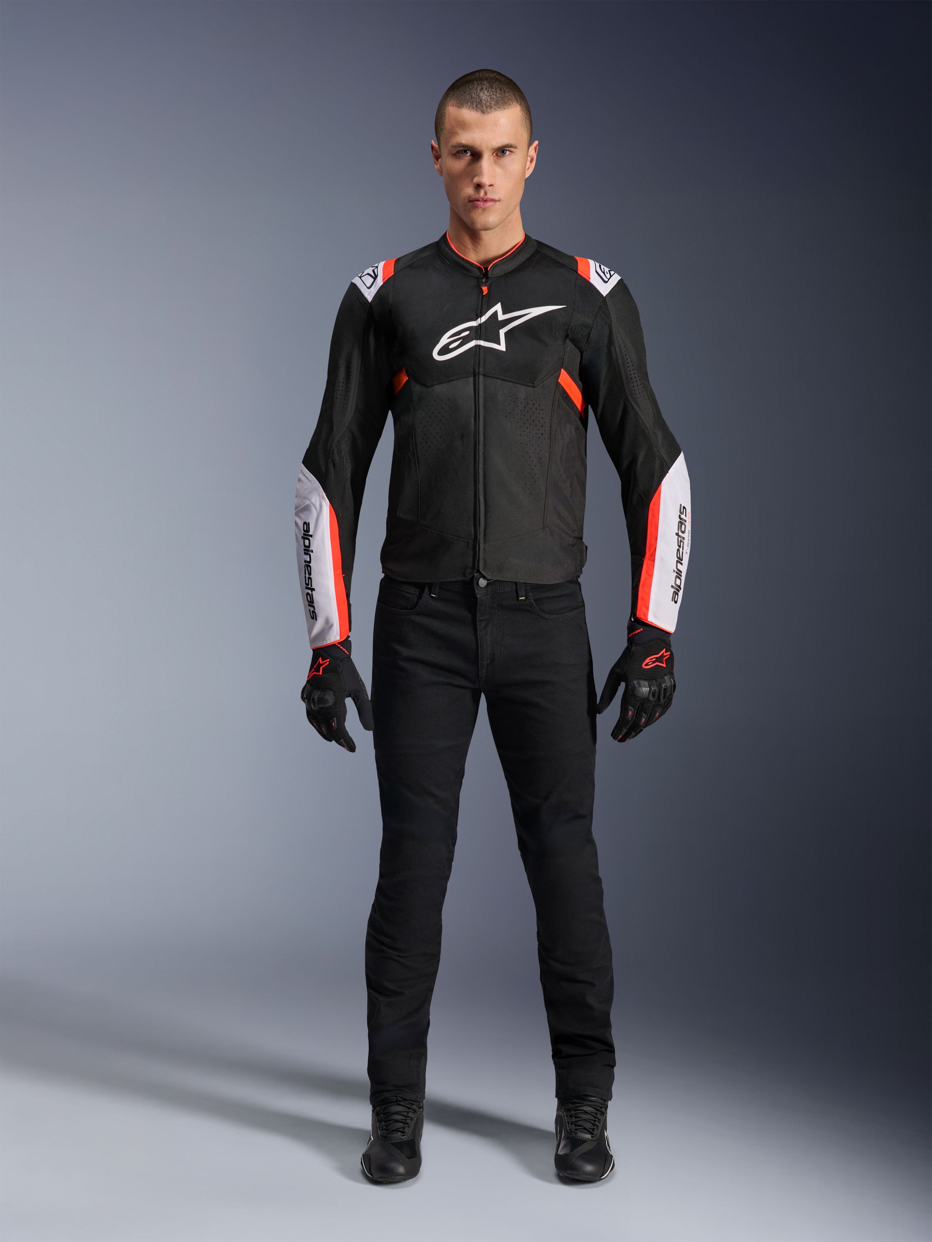Mandlig model iført Alpinestars T-Sps Air V2 jakke i sort, hvid og rød fluo, med store tech-mesh-paneler og tydelig hvid branding på brystet og ærmerne, kombineret med sorte tekniske kørebukser og handsker, urban sport motorcykeludstyr