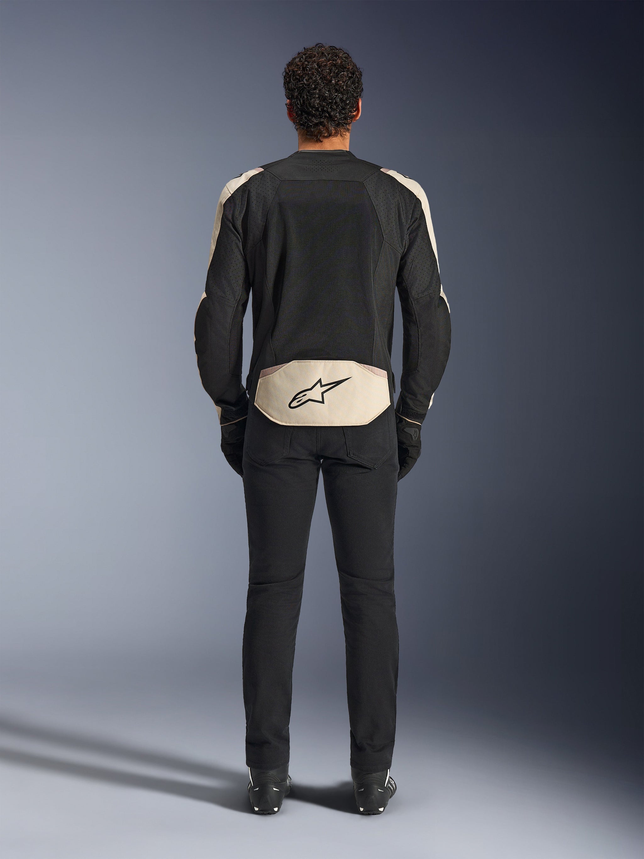 Alpinestars T-SPS Air V2-jakke, set bagfra, Black Mastic Aluminum, tekstil motorcykeljakke med store mesh-paneler til ventilation, lysebeige detaljer på skuldre og lænd med Alpinestars-logoet, vist med sorte kørebukser og støvler