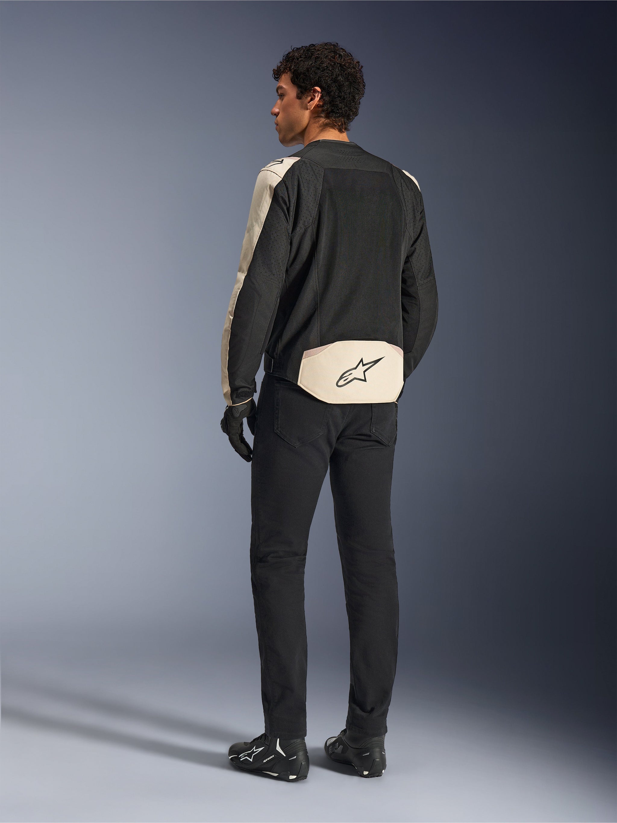 Alpinestars T-SPS Air V2 jakke, tekstil motorcykeljakke, Black Mastic Aluminum, set bagfra på model, med omfattende tech mesh-paneler for optimal luftgennemstrømning, beige detaljer på ærmer og lænd med Alpinestars-logo, kombineret med sorte bukser og støvler.