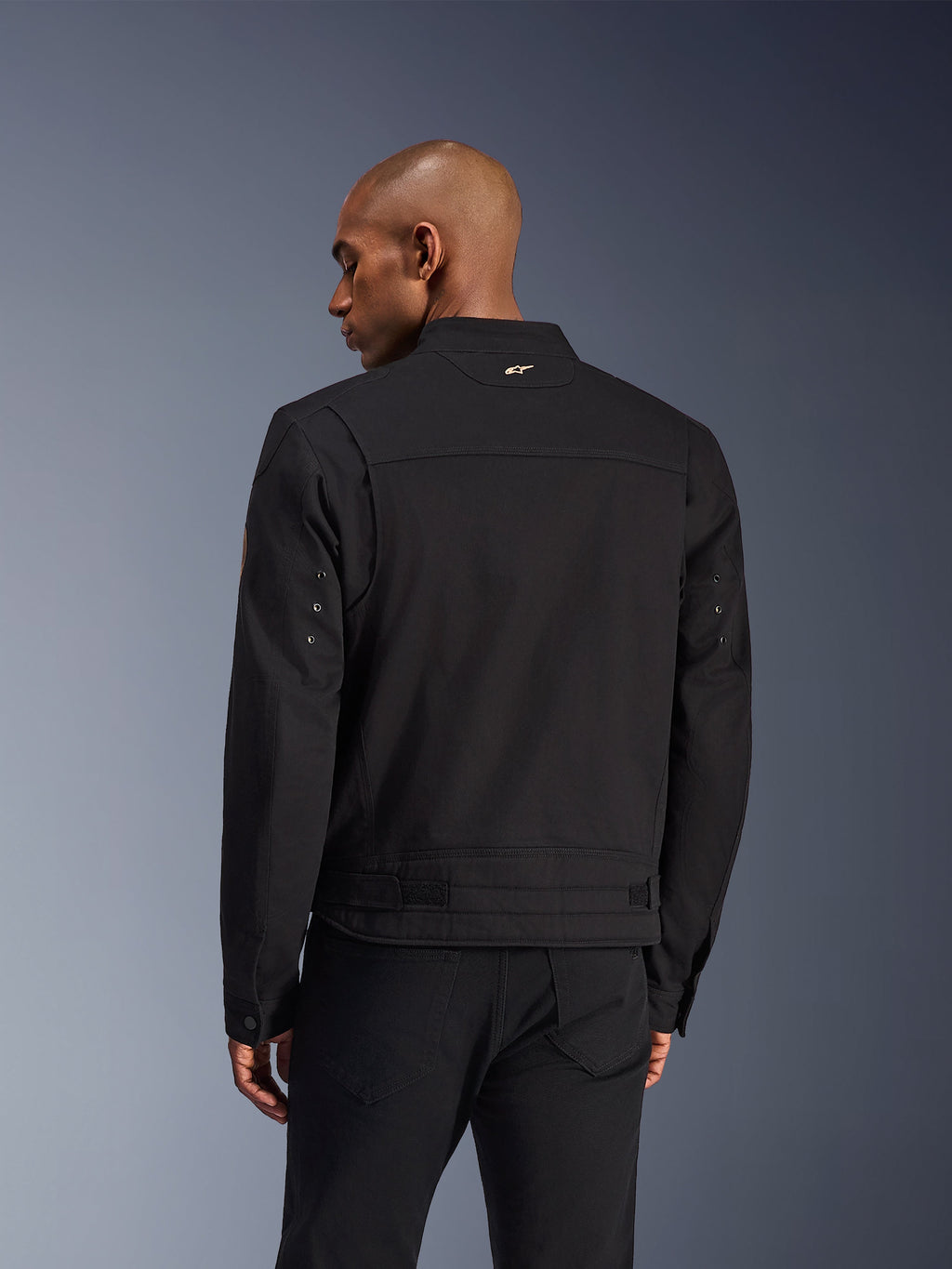 Alpinestars Bruiser jakke, sort urban motorcykeljakke, set bagfra, med et arbejdstøjsinspireret design, lille guldlogo på kraven og justerbare taljestropper, slidstærk tekstilkonstruktion til bykørsel