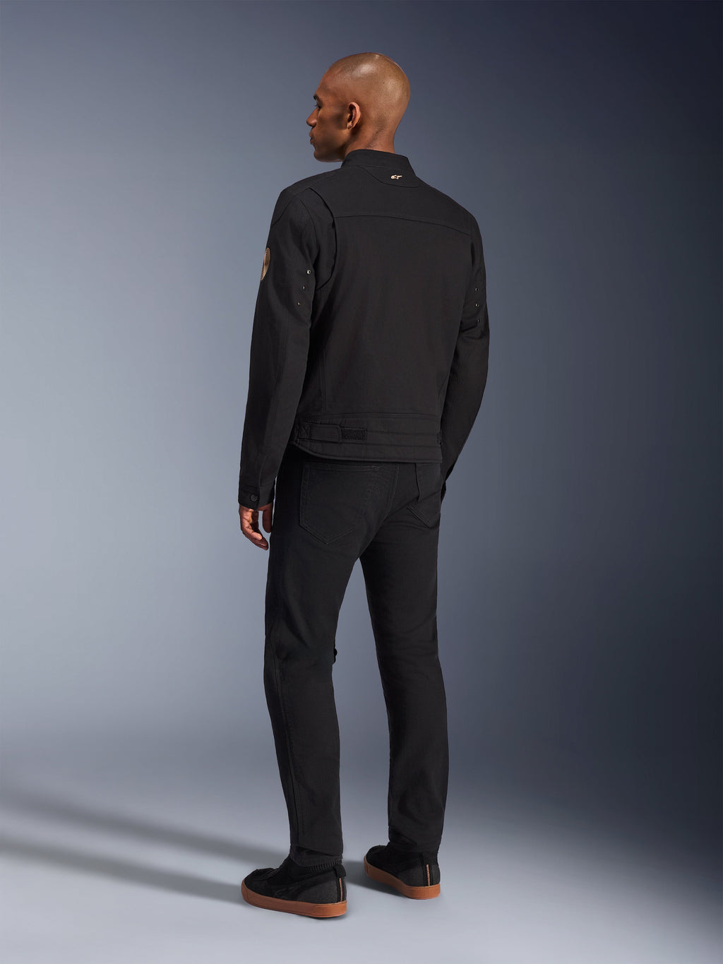 Alpinestars Bruiser-jakke, urban sort motorcykeljakke, set bagfra på en model, klassisk arbejdstøjsstil, med plisserede skulderpaneler og Oscar-spiralmærke på ærmet, båret med sorte bukser og afslappede sorte sko med gummisåler.
