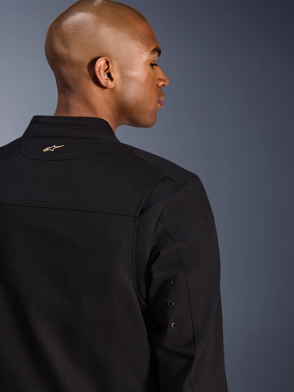 Alpinestars Bruiser jakke, urban motorcykeljakke i tekstil, sort, nærbillede af ryggen med broderet guldlogo, detaljer med metaløjer på ærmet og plisserede skulderpaneler for øget mobilitet