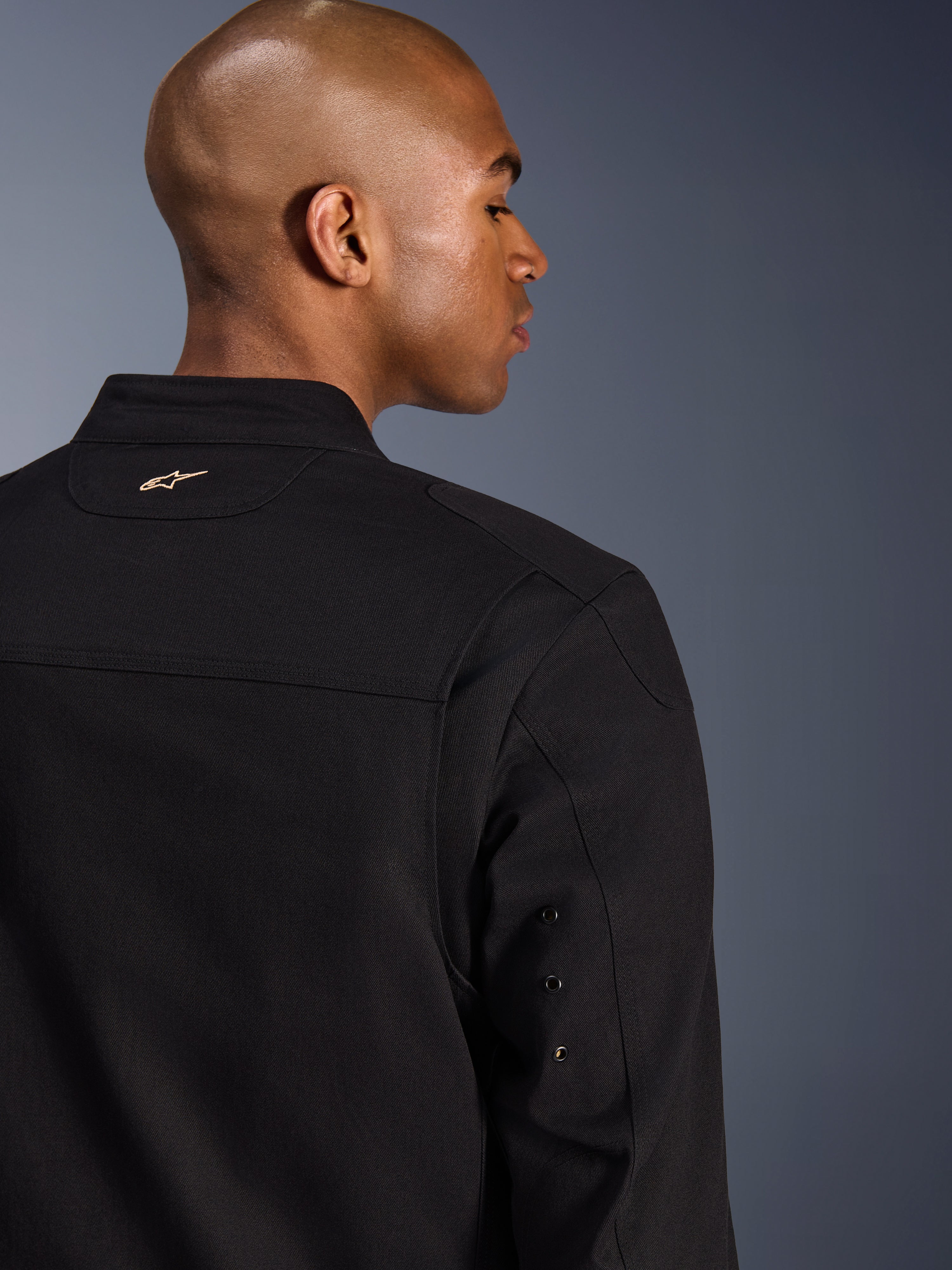 Alpinestars Bruiser jakke, urban motorcykeljakke i tekstil, sort, nærbillede af ryggen med broderet guldlogo, detaljer med metaløjer på ærmet og plisserede skulderpaneler for øget mobilitet