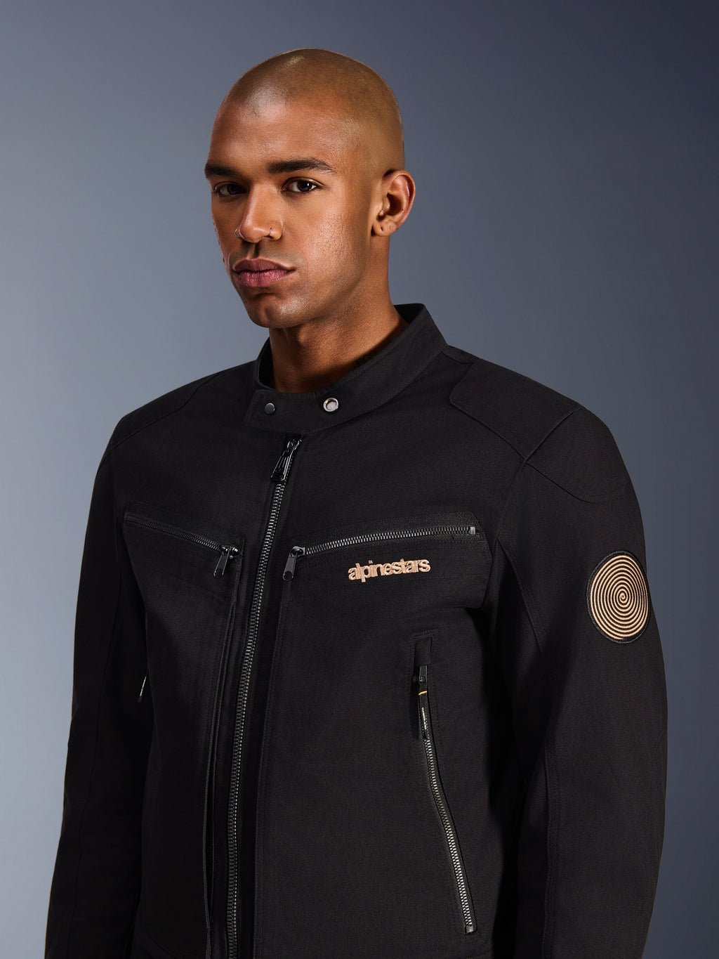 Mandlig model iført Alpinestars Bruiser Jakke, sort, urban motorcykeljakke i polycotton twill, med lynlåslommer, guldbroderet logo på brystet og Oscar-spiral-patch på ærmet, moderne pendlestil
