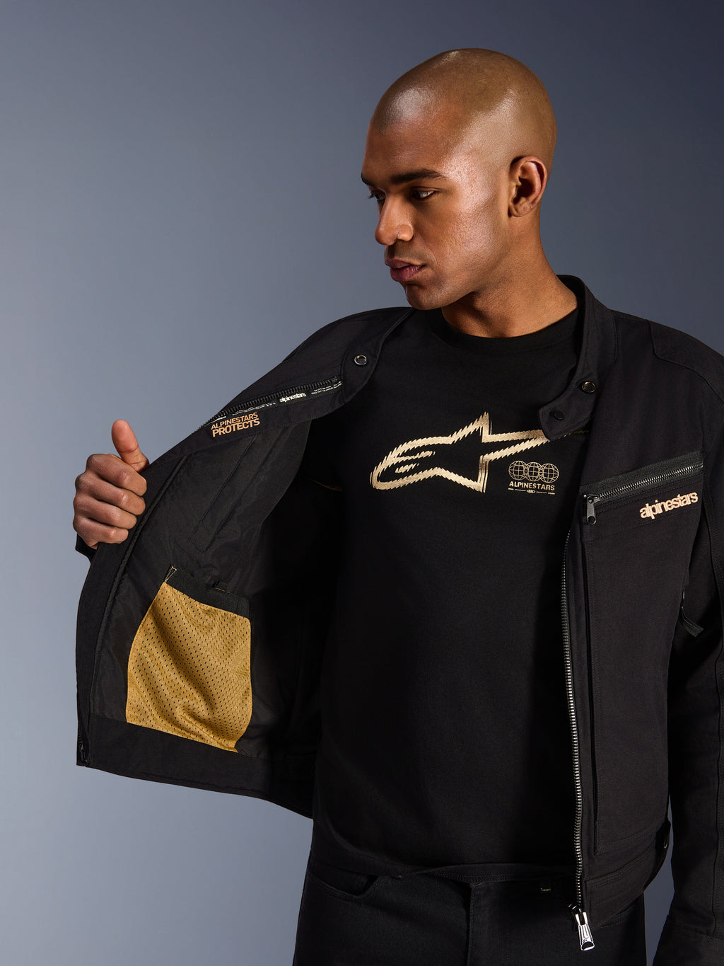 Alpinestars Bruiser jakke, sort urban motorcykeljakke, nærbillede der viser det åbne interiør med guld mesh-lomme og 'Alpinestars Protects' branding, båret over en sort t-shirt med logo, tekstilkørselsudstyr med workwear-æstetik