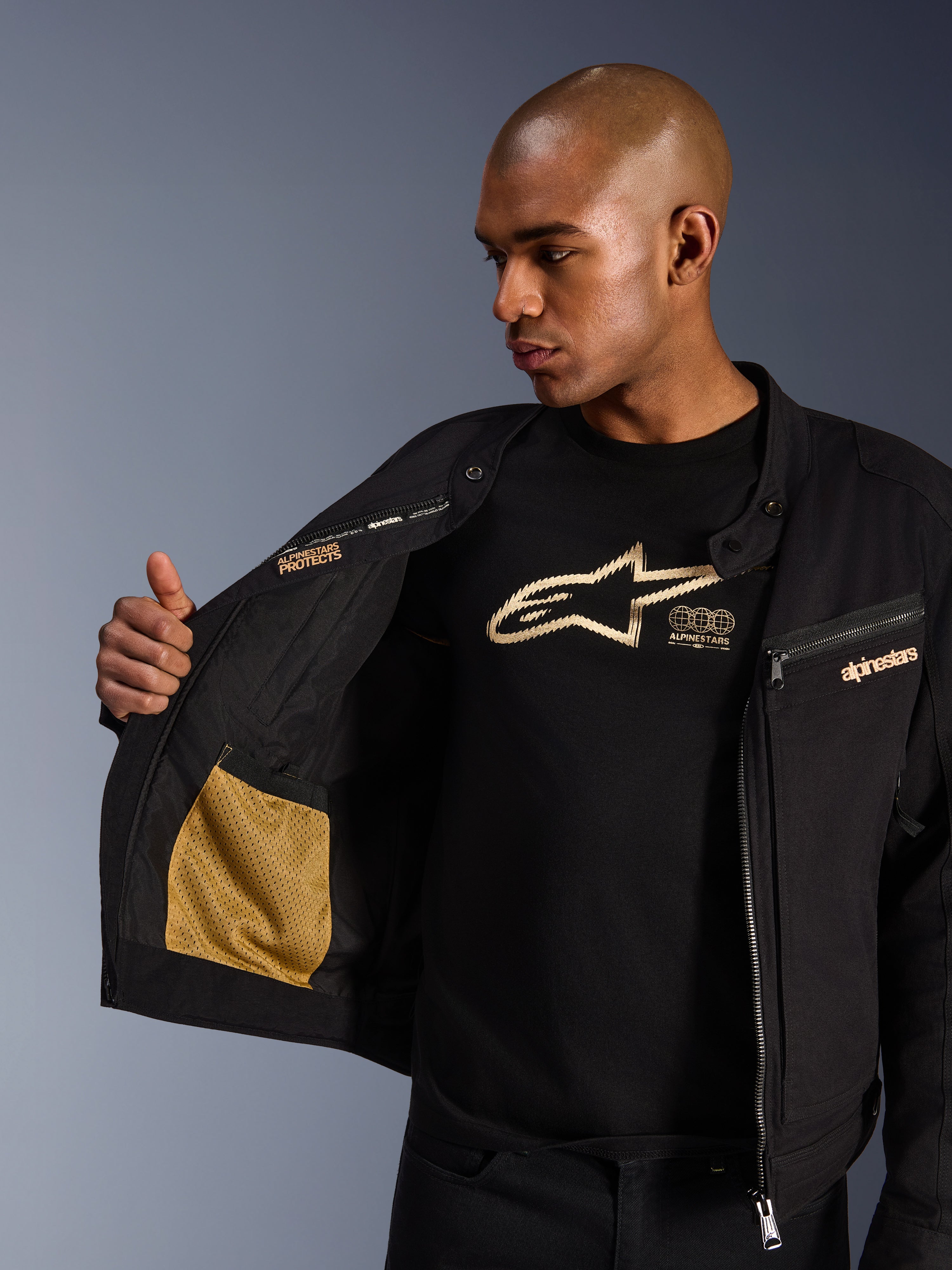 Alpinestars Bruiser jakke, sort urban motorcykeljakke, nærbillede der viser det åbne interiør med guld mesh-lomme og 'Alpinestars Protects' branding, båret over en sort t-shirt med logo, tekstilkørselsudstyr med workwear-æstetik