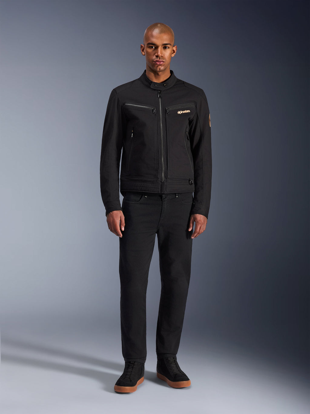 Alpinestars Bruiser jakke, sort urban motorcykeljakke, båret af model med sorte bukser og high-top sneakers, set forfra med brystlommer med lynlås og broderet logo, klassisk arbejdstøjsinspireret design til daglig pendling