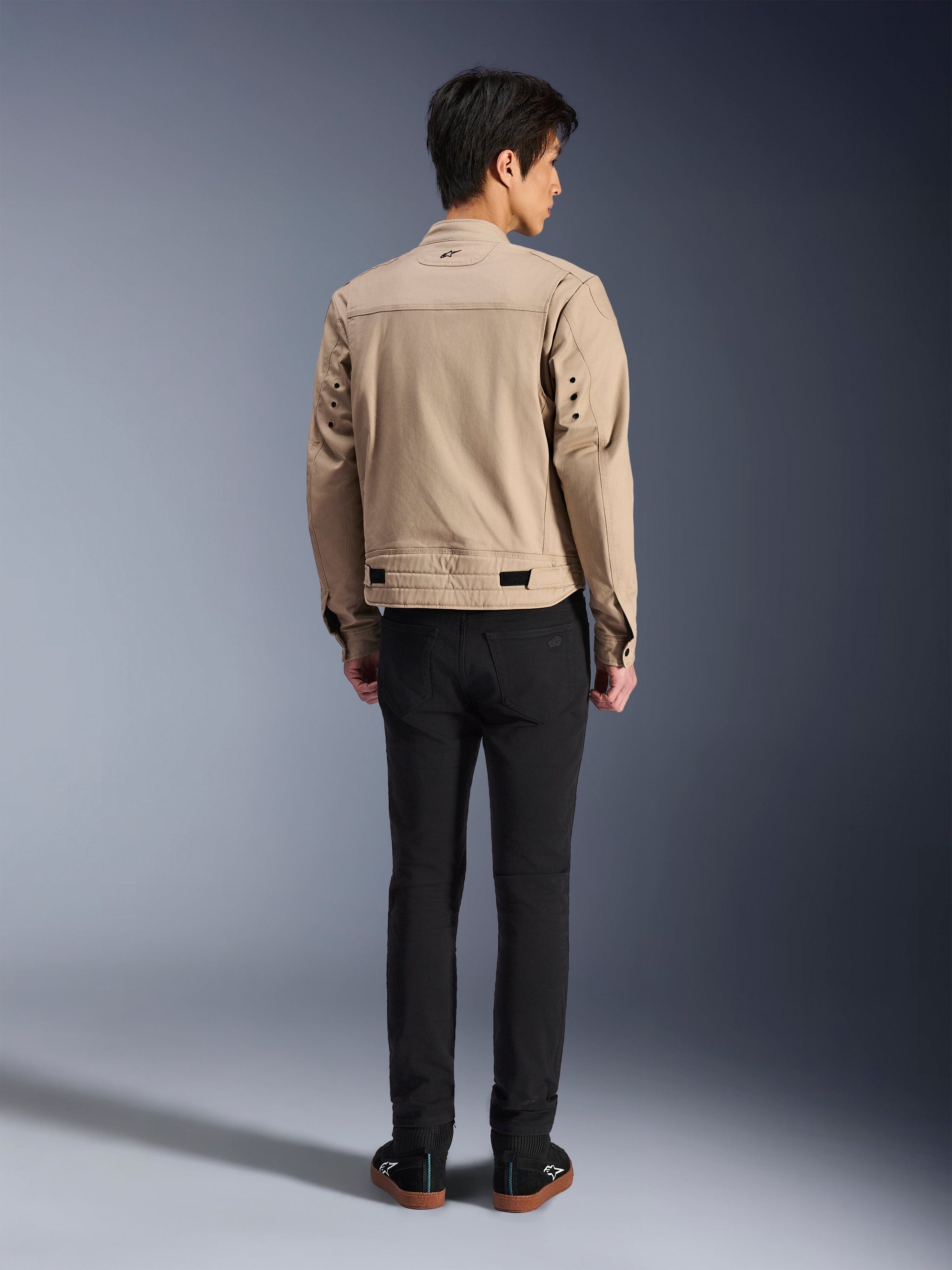 Alpinestars Bruiser Jacket i brun, urban motorcykeljakke set bagfra på model, arbejdstøjsinspireret design med metaløskner på ryggen og taljejusteringer, kombineret med sorte bukser og sneakers, moderne street-kørestil.