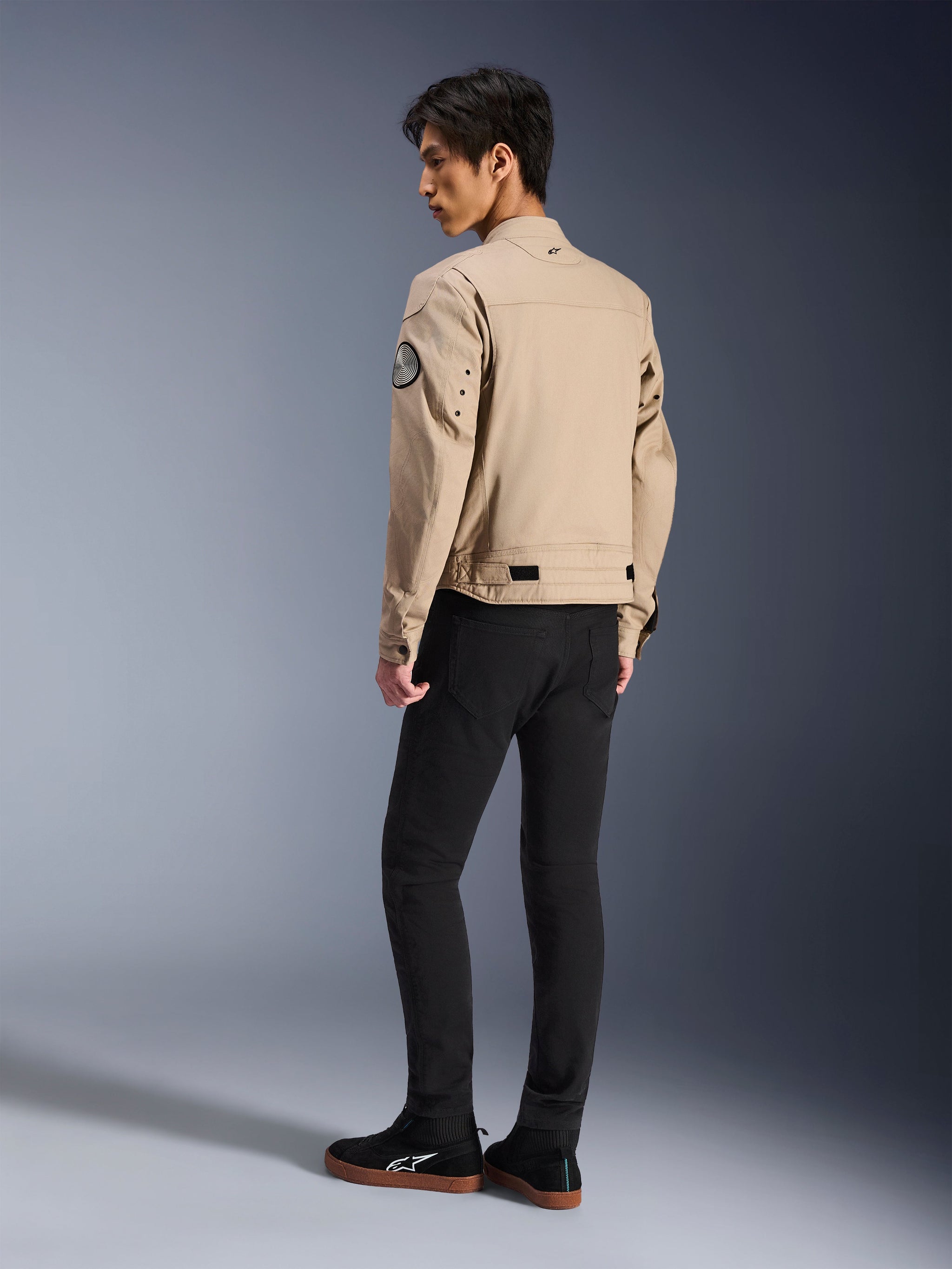 Mandlig model set bagfra iført Alpinestars Bruiser-jakke i tan, urban moderne motorcykeljakke med arbejdstøjs-styling, med et rundt ærmemærke, justeringsremme i taljen og artikulerede skuldre, kombineret med sorte bukser og sorte sneakers.