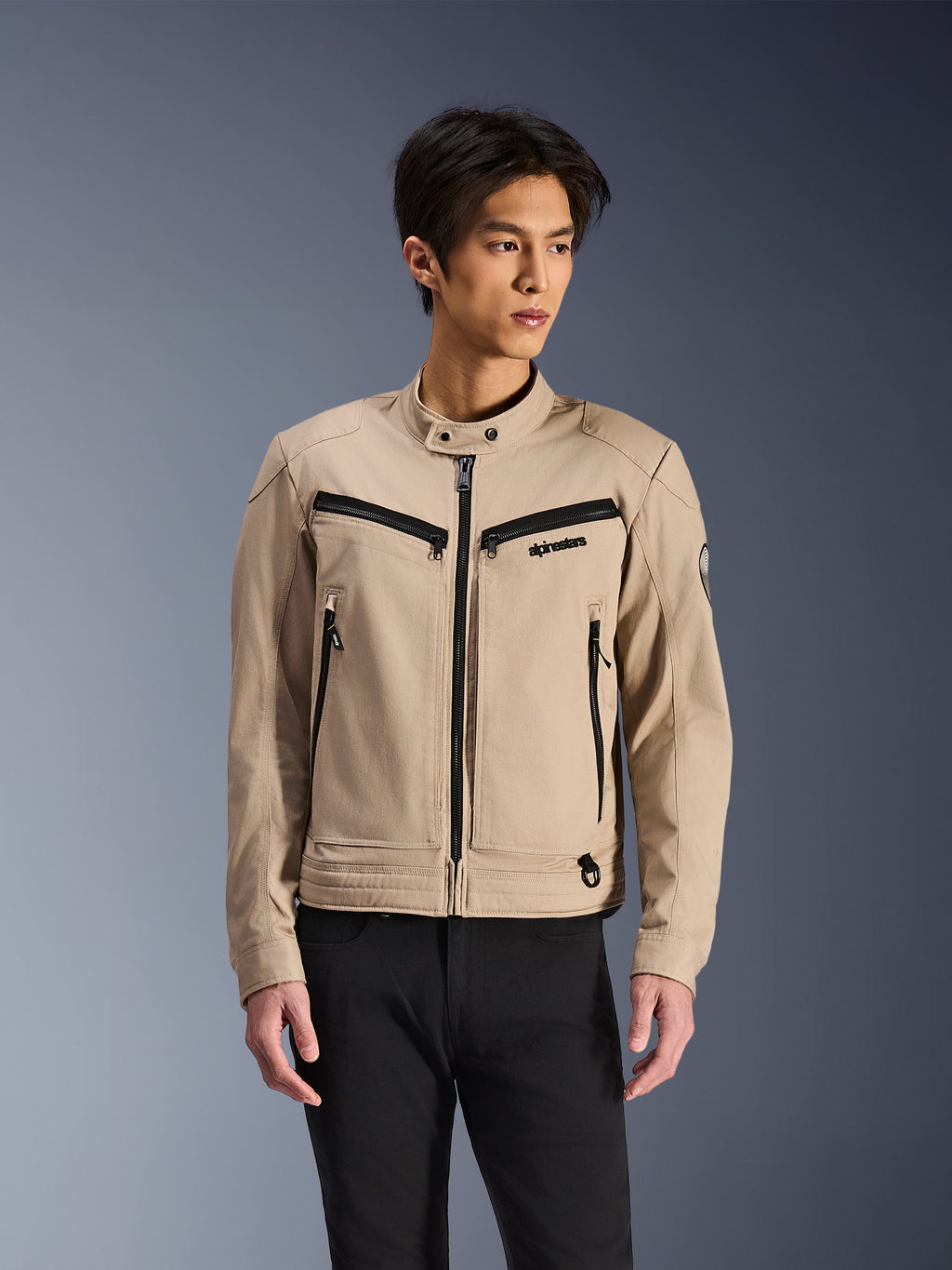 Alpinestars Bruiser Jacket, beige motorcykeljakke båret af mandlig model, med sorte lynlåsdetaljer på bryst- og sidelommer, høj krave med trykknapper, arbejdstøj-inspireret urbant køreudstyr, kombineret med sorte bukser
