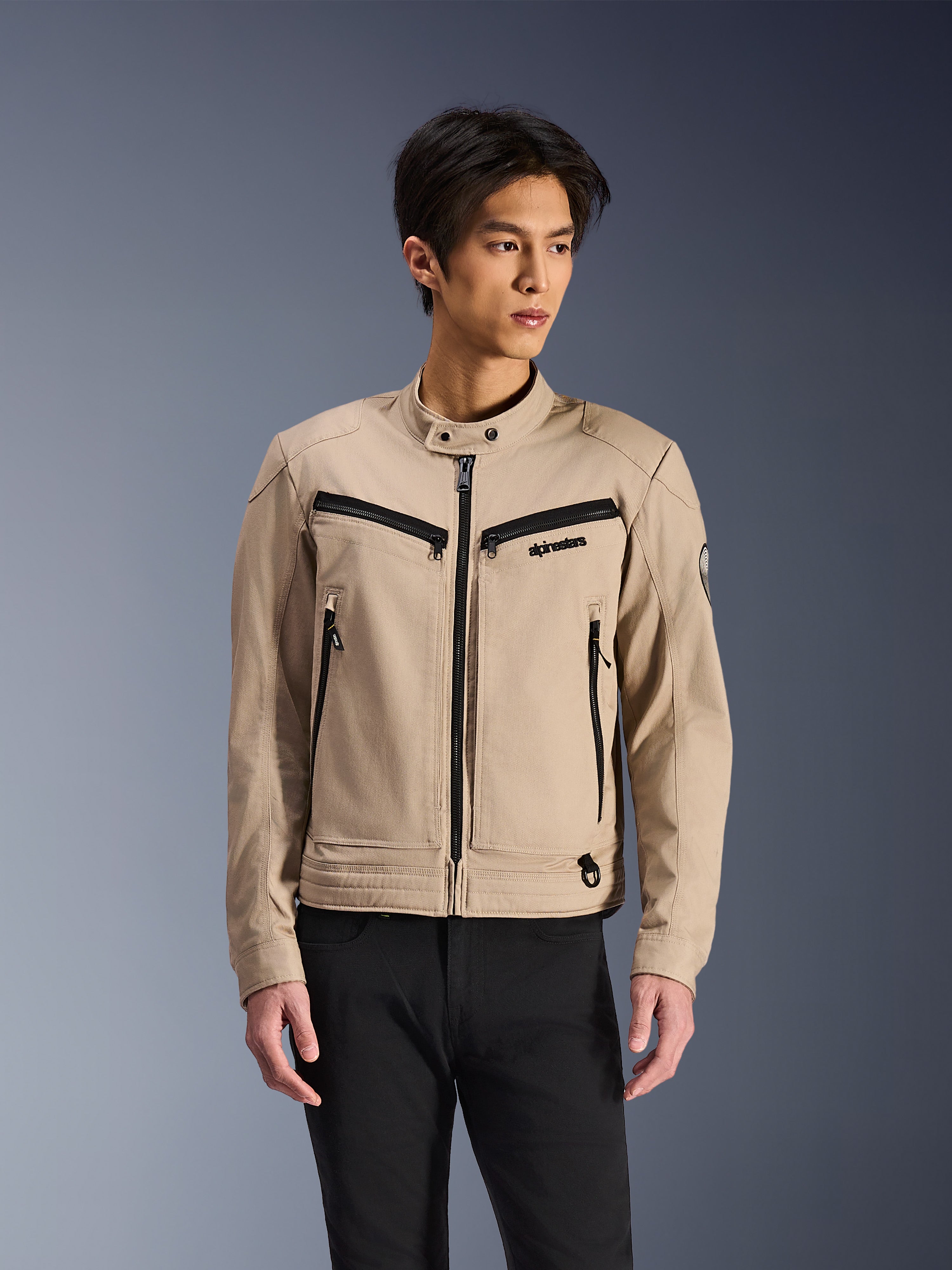 Alpinestars Bruiser Jacket, beige motorcykeljakke båret af mandlig model, med sorte lynlåsdetaljer på bryst- og sidelommer, høj krave med trykknapper, arbejdstøj-inspireret urbant køreudstyr, kombineret med sorte bukser