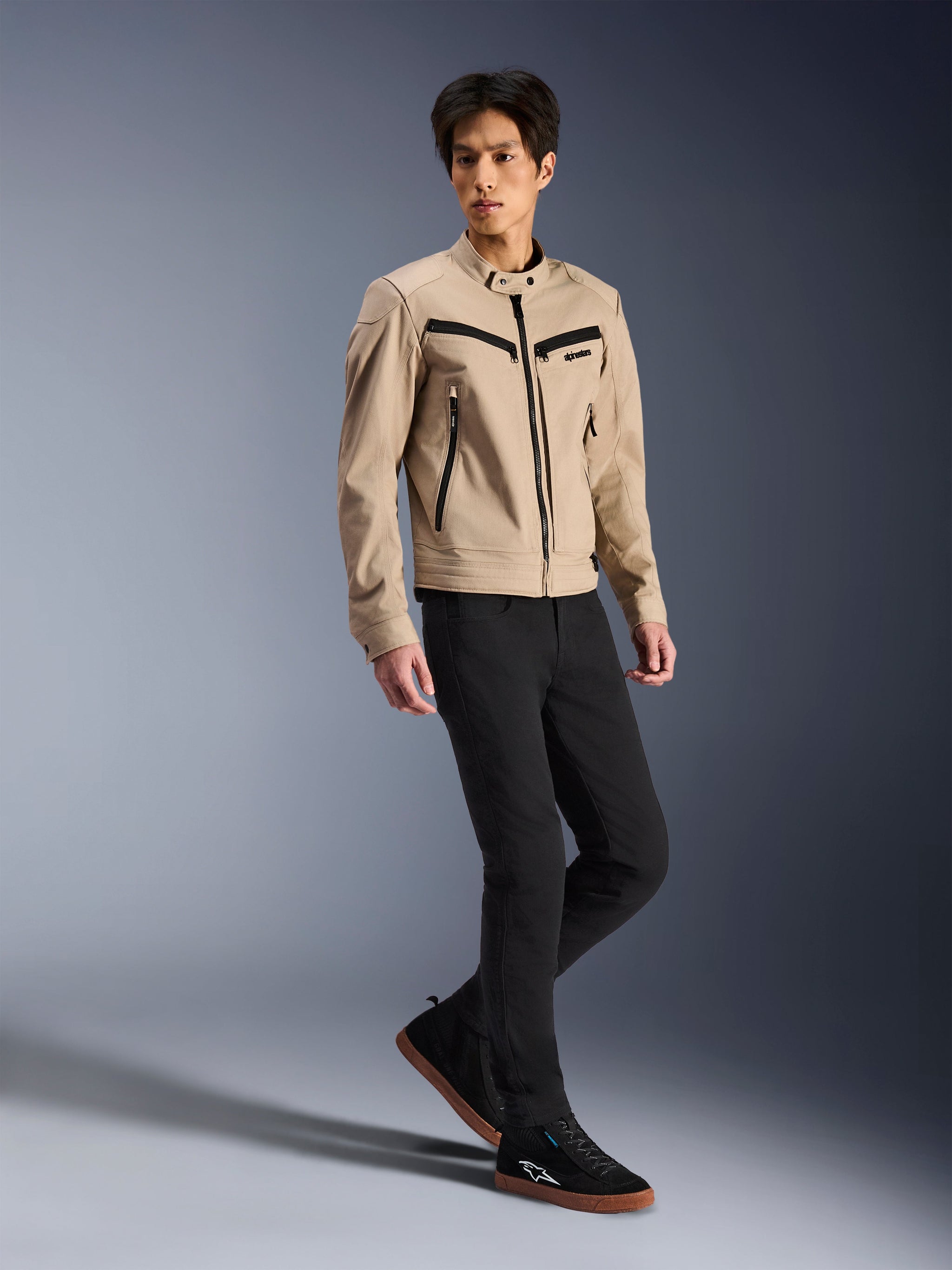 Alpinestars Bruiser-jakke, urban motorcykeljakke, tan, båret af en mandlig model med sorte slim-fit bukser og sorte sneakers, arbejdstøjsinspireret tekstilkonstruktion med sorte lynlåsdetaljer, moderne urban street-stil.