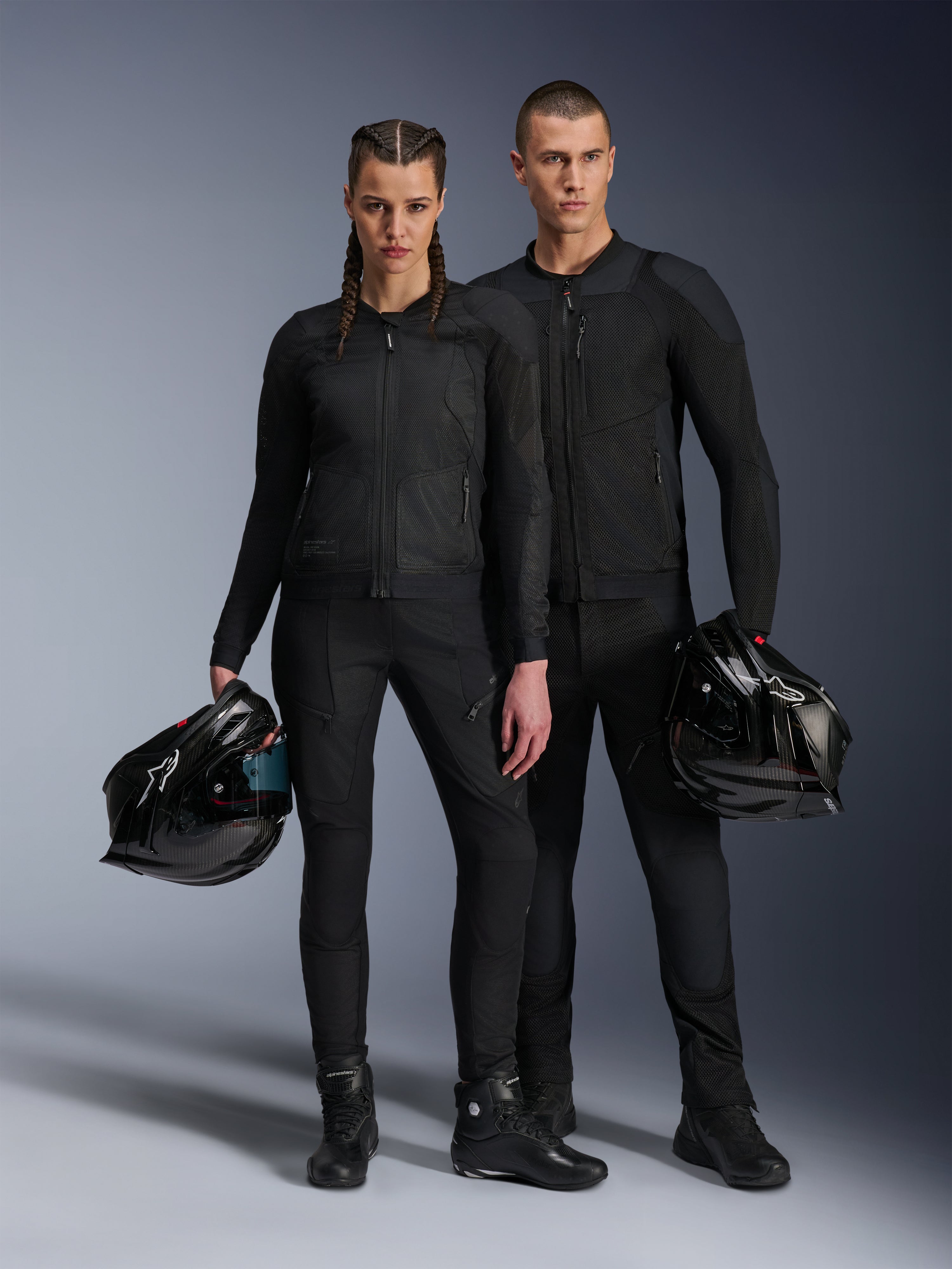 Mandlig og kvindelig model iført sorte Alpinestars Troop-Air Pro-jakker, teknisk mesh-køreudstyr kombineret med matchende sorte bukser og støvler, hver med en sort hjelm, der fremviser koordineret urbant motorcykeltøj.