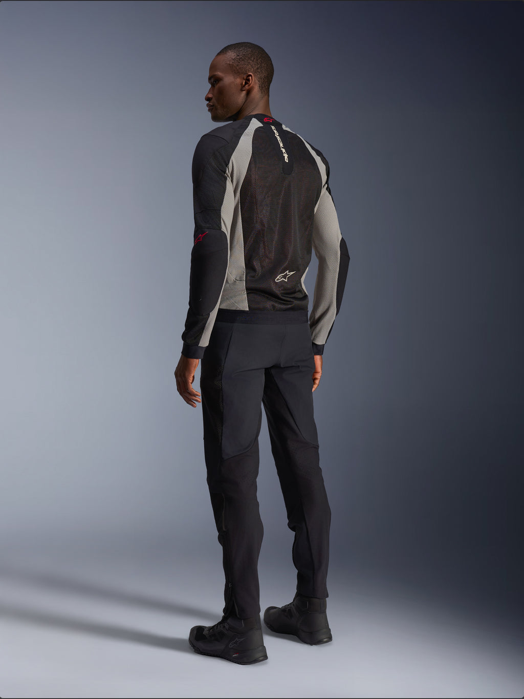 Model wearing Alpinestars Troop-Air Jacket from the back, Black Ash Gray and Bright Red accents, technical mesh construction for ventilation, paired with black motorcycle pants and riding boots, urban contemporary style