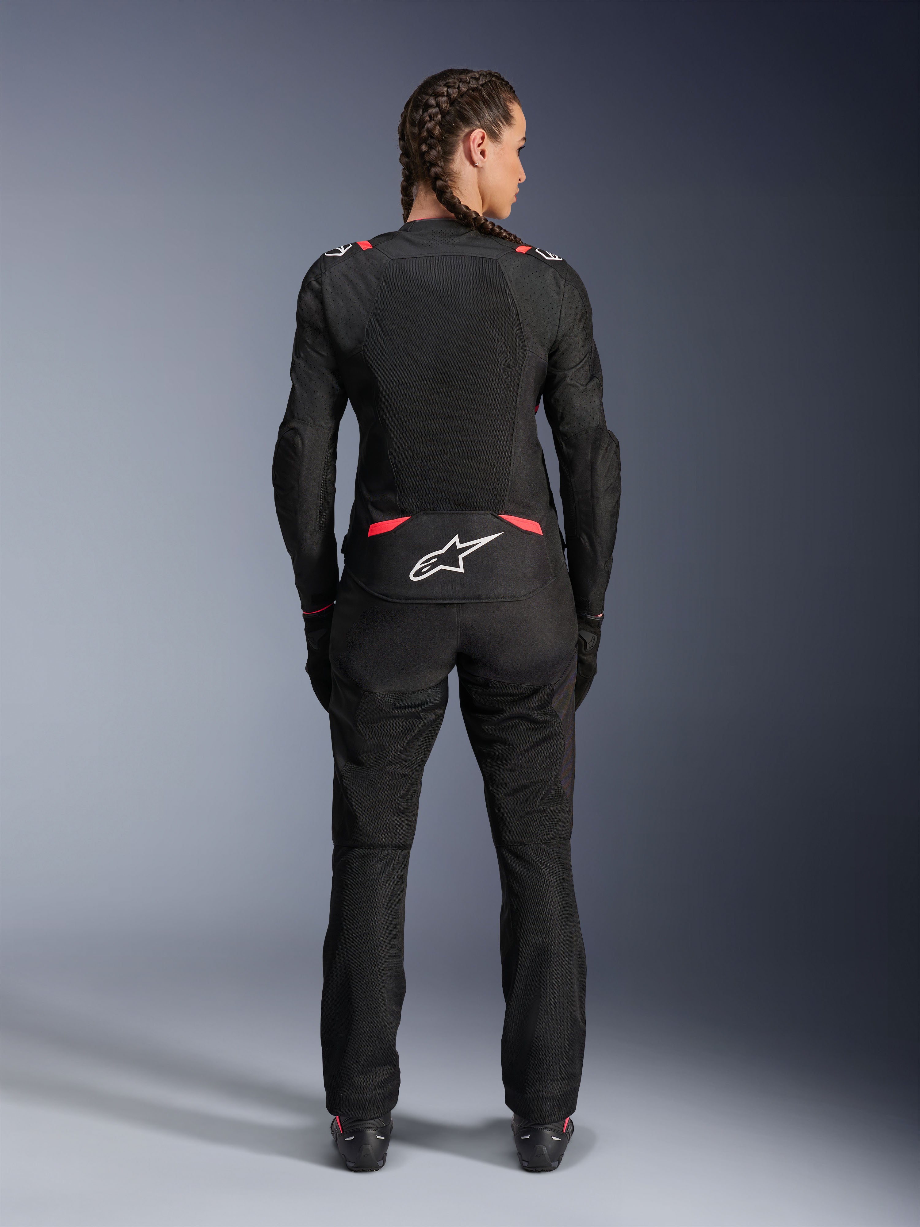 Alpinestars Stella T-SPS Air V2-jakke, motorcykeljakke i tekstil til kvinder, sort og Diva Pink, set bagfra på model med flettet hår, med store mesh-paneler til ventilation, hvidt Astars-logo på lænden, kombineret med matchende sorte kørebukser.