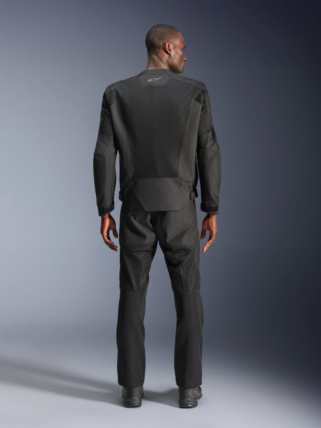 Set bagfra af en mand iført Alpinestars C-1 Air Jacket i sort, urban motorcykeljakke med tech mesh-paneler, matchende sorte tekstilbukser og støvler, rent æstetisk design med reflekterende logo øverst på ryggen