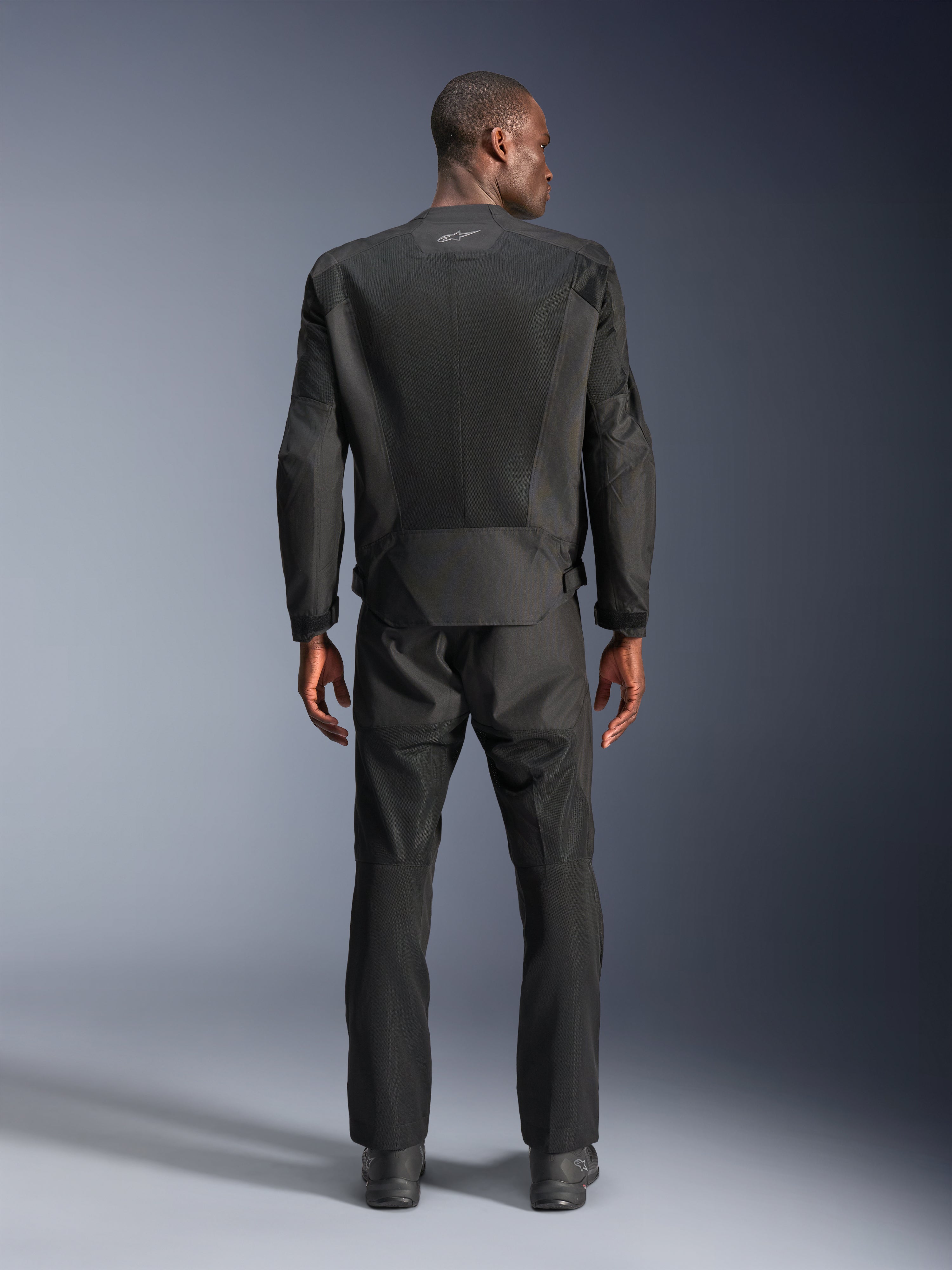Set bagfra af en mand iført Alpinestars C-1 Air Jacket i sort, urban motorcykeljakke med tech mesh-paneler, matchende sorte tekstilbukser og støvler, rent æstetisk design med reflekterende logo øverst på ryggen