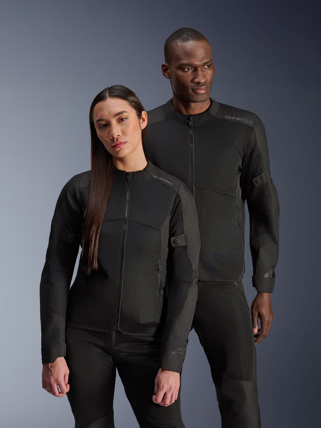 En mand og en kvinde iført Alpinestars C-1 Air Jacket og Stella C-1 Air Jacket, matchende sorte urbane tekstilmotorcykeljakker med mesh-paneler til ventilation, kombineret med sorte kørebukser for et rent og moderne look.