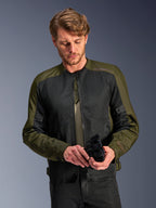 Mand iført Alpinestars C-1 Air-jakken i Khaki, urban motorcykeljakke med olivengrønt tekstil og sorte mesh-paneler, kigger ned mens han holder sorte motorcykelhandsker, rent og moderne design til landevejskørsel.