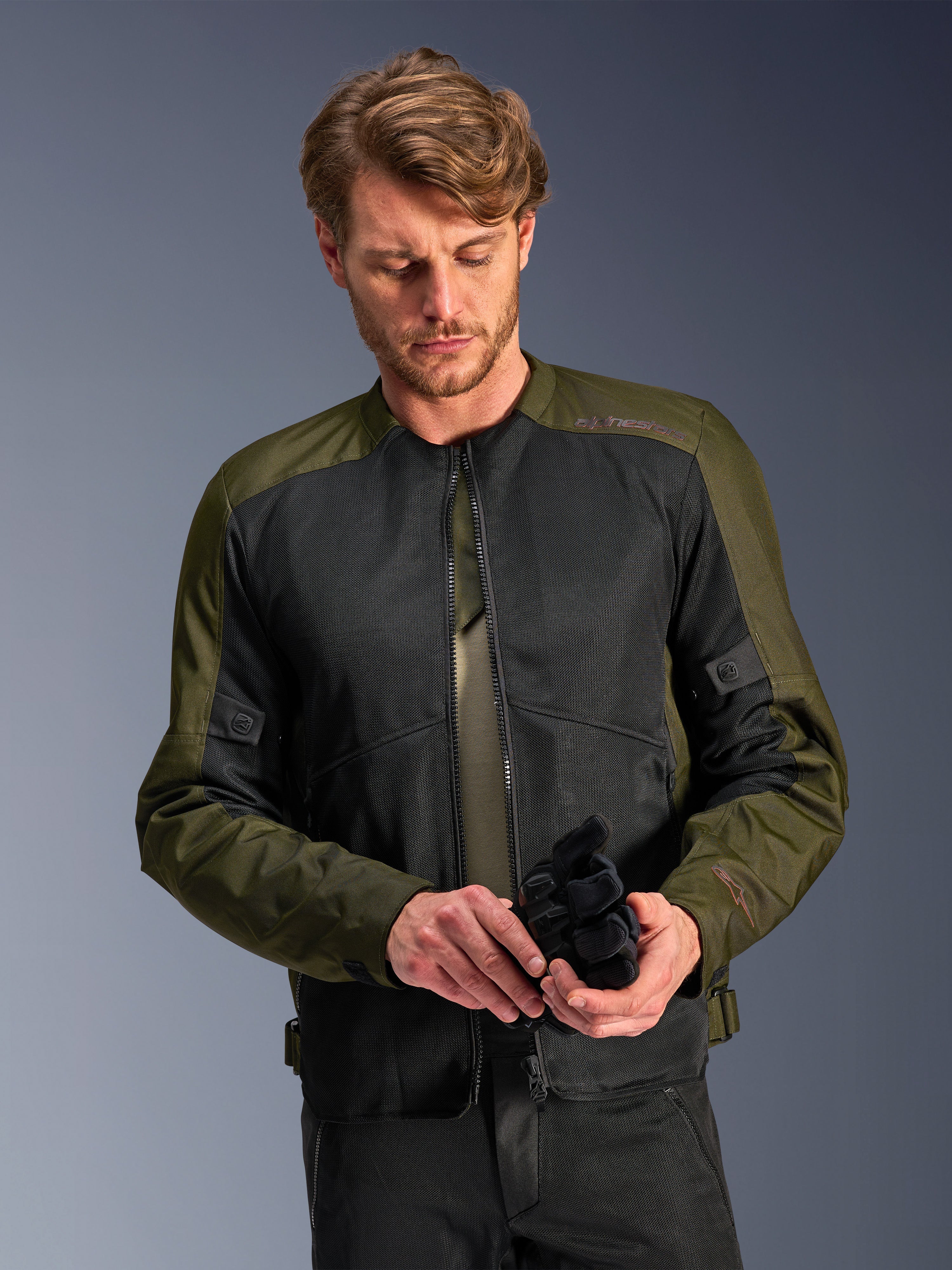 Mand iført Alpinestars C-1 Air-jakken i Khaki, urban motorcykeljakke med olivengrønt tekstil og sorte mesh-paneler, kigger ned mens han holder sorte motorcykelhandsker, rent og moderne design til landevejskørsel.