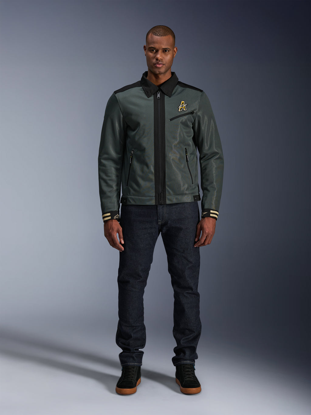 Alpinestars Ignite Air Jakke i Urban Gray Black Blue, motorcykeljakke i Harrington-stil med skjortekrave og mesh-konstruktion, båret af en mand med mørke denimjeans og sorte sneakers, urban streetwear til motorcyklister.