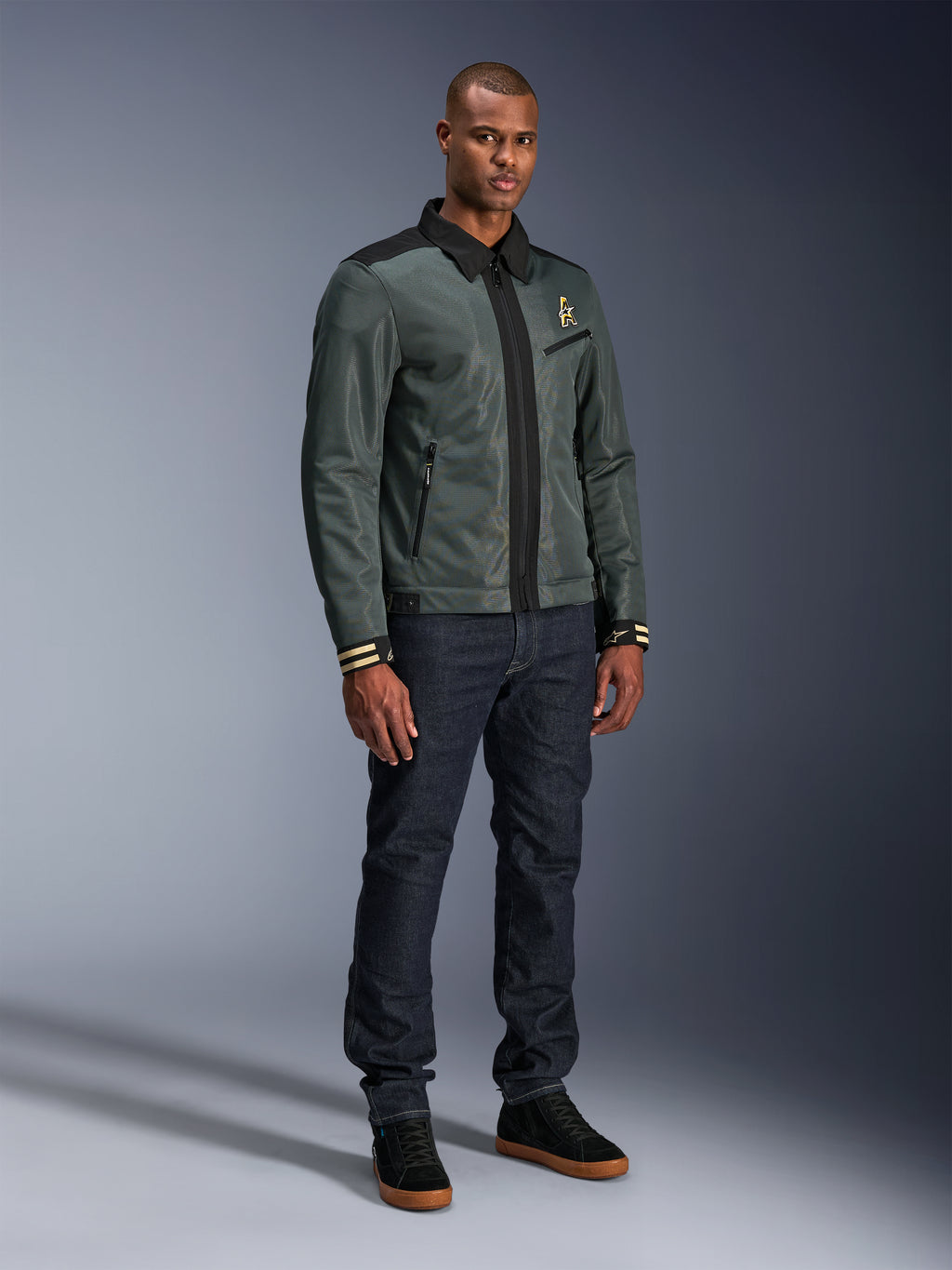 Alpinestars Ignite Air-jakke, Urban Gray Black Blue, urban motorcykeljakke, båret af en model med mørke denimjeans og sorte sneakers, med mesh-krop, traditionel skjortekrave, Harrington-inspireret silhuet og guldfarvet Alpinestars-branding.