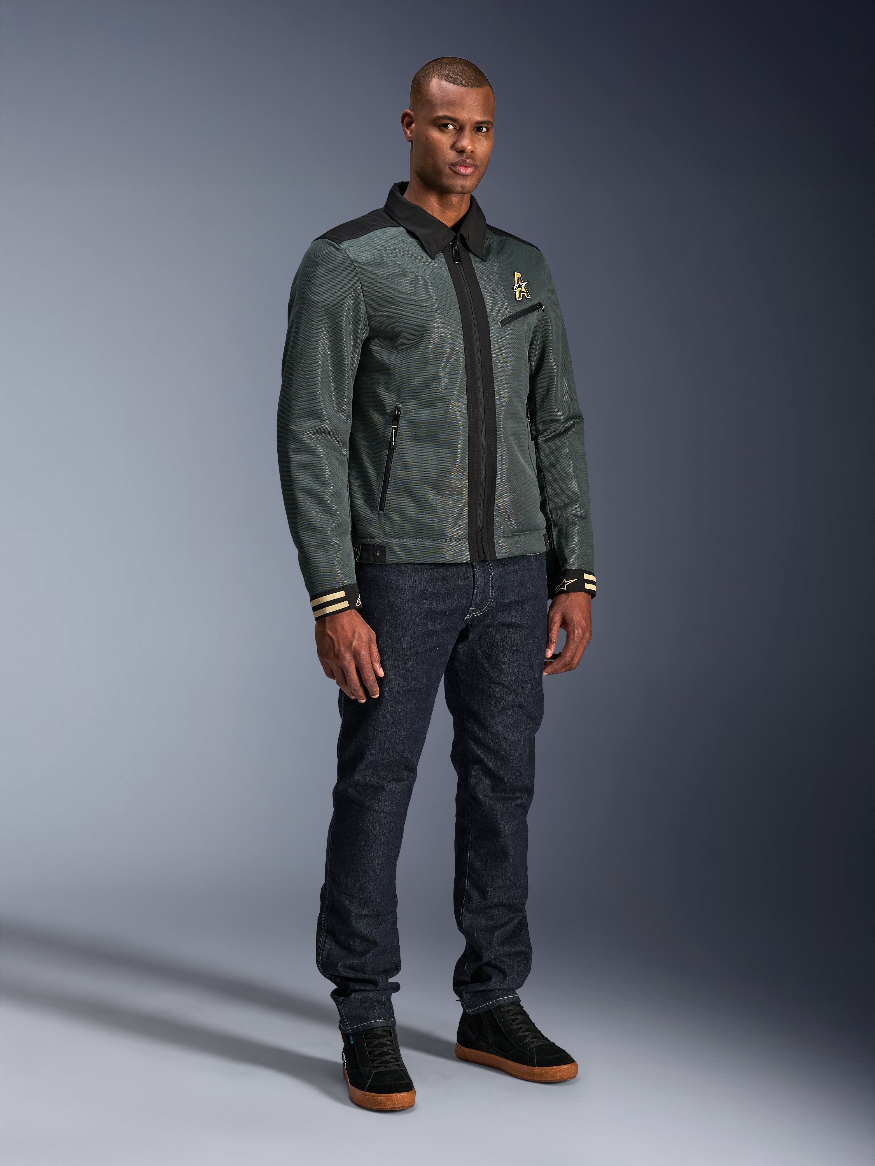 Alpinestars Ignite Air-jakke, Urban Gray Black Blue, urban motorcykeljakke, båret af en model med mørke denimjeans og sorte sneakers, med mesh-krop, traditionel skjortekrave, Harrington-inspireret silhuet og guldfarvet Alpinestars-branding.
