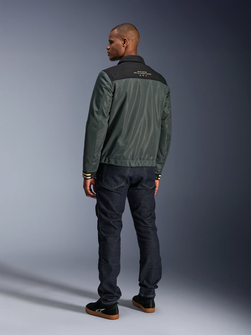 Alpinestars Ignite Air Jakke i Urban Gray Black Blue, set bagfra på model, med mesh-krop, sort skulderstykke og hvid tekst, stribede manchetter, kombineret med mørke denimjeans og sorte sneakers, urban livsstils-motorcykelbeklædning