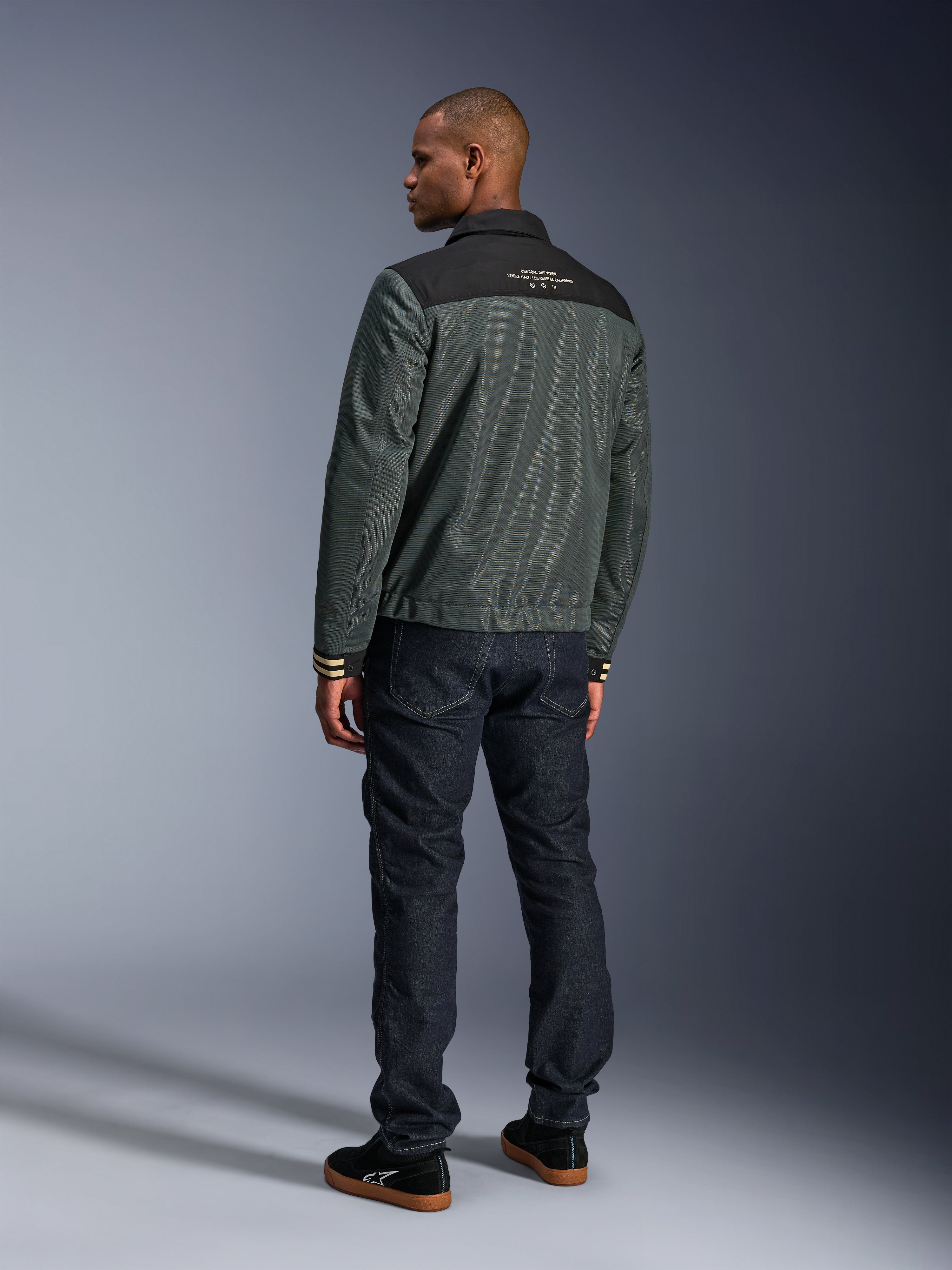Alpinestars Ignite Air Jakke i Urban Gray Black Blue, set bagfra på model, med mesh-krop, sort skulderstykke og hvid tekst, stribede manchetter, kombineret med mørke denimjeans og sorte sneakers, urban livsstils-motorcykelbeklædning