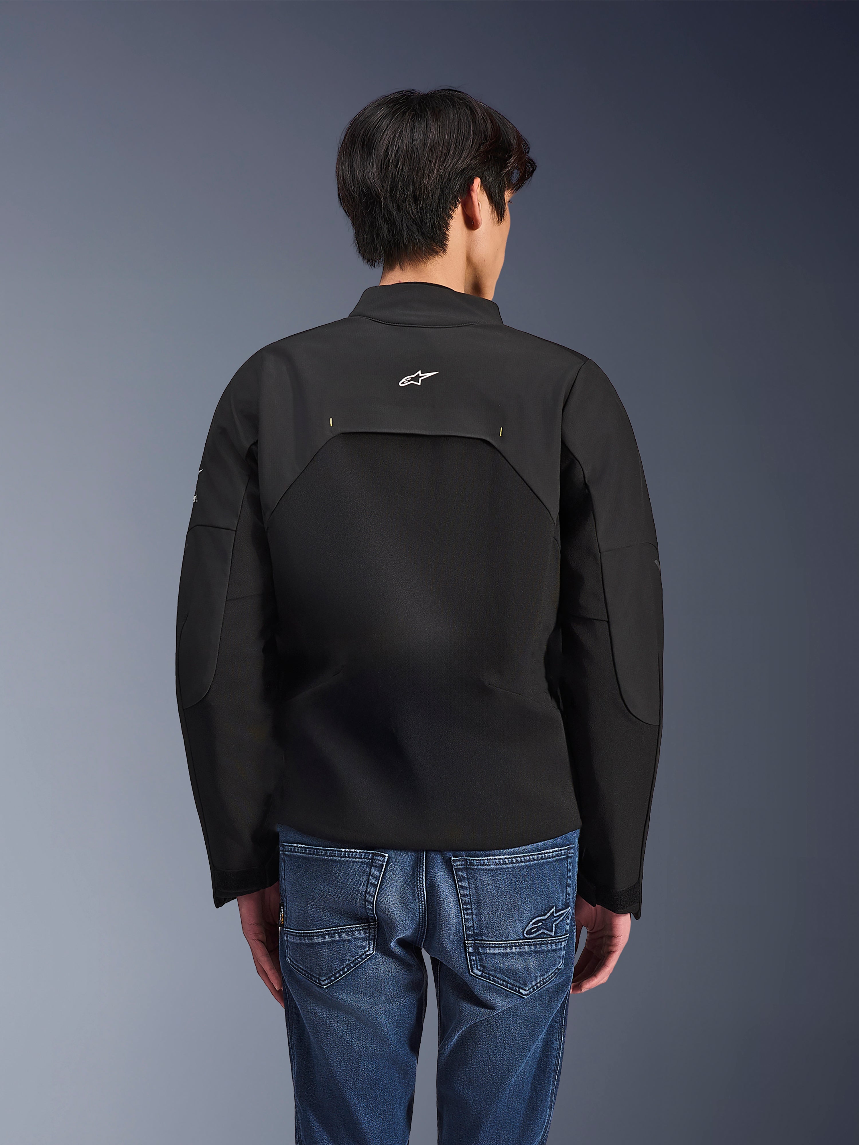 Alpinestars Provoke-jakke, urban motorcykeljakke, sort, set bagfra med hvidt Astars-logo og tekniske ripstop-paneler, båret med blå denimjeans, moderne design til pendling i byen