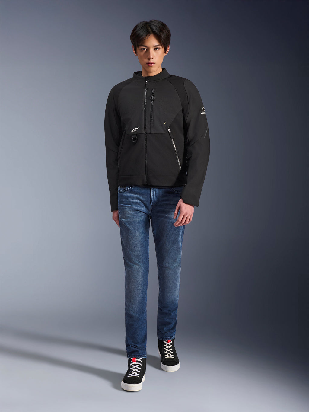 Mandlig model iført Alpinestars Provoke Jacket i sort, urban tekstil motorcykeljakke med tekniske lynlåse og ripstop softshell-paneler, kombineret med blå jeans og sorte sneakers, moderne urban rider-stil