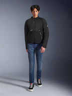 Mandlig model iført Alpinestars Provoke Jacket i sort, urban tekstil motorcykeljakke med tekniske lynlåse og ripstop softshell-paneler, kombineret med blå jeans og sorte sneakers, moderne urban rider-stil